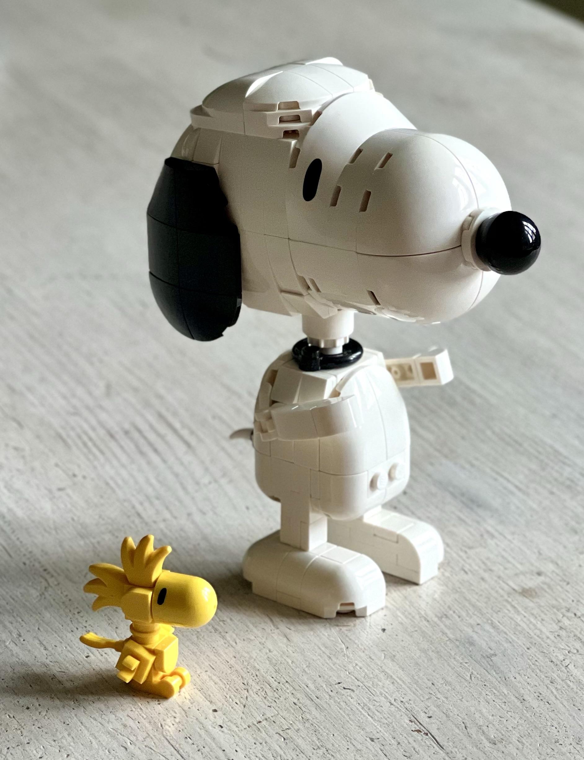 A large white and black LEGO model of Snoopy stands behind a smaller model of Woodstock. They are placed on a white-ish, shabby wooden table.