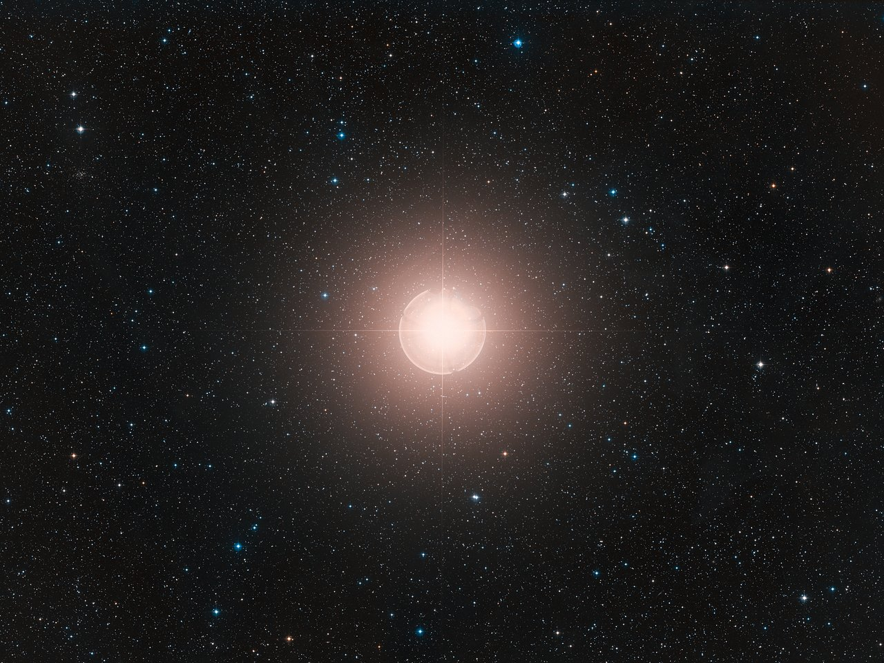 A bright orange-red star is visible at the centre of the image. A broad halo fades into the surrounding darkness. Many stars are scattered across the field. 