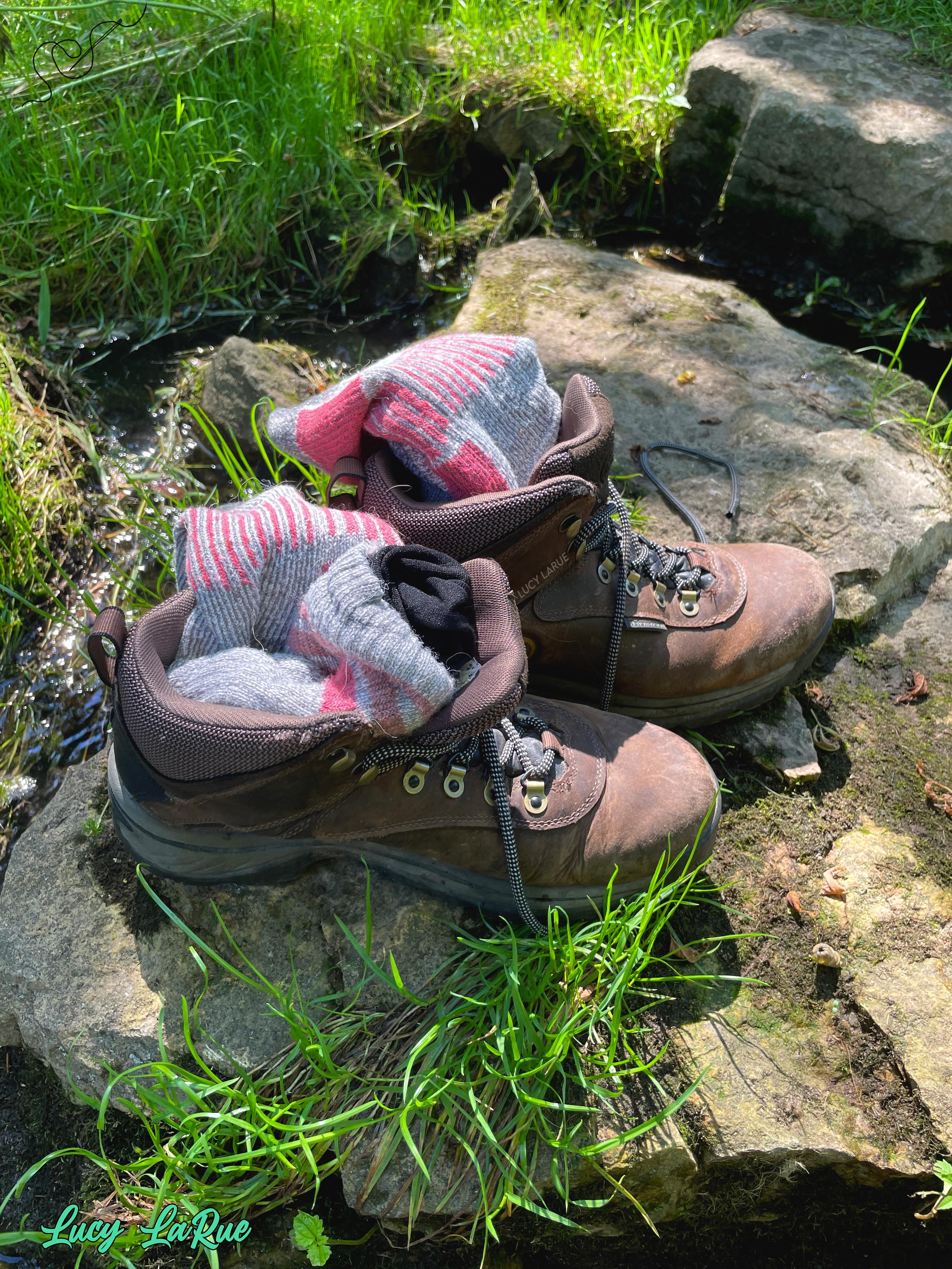A pair of brown hiking boots resting on a rock covered in mosses with a stream and bright green grasses surrounding it, each shoe stuffed with a grey and pink woolen hiking sock. Their owner has gone barefoot in the sparkling creek.