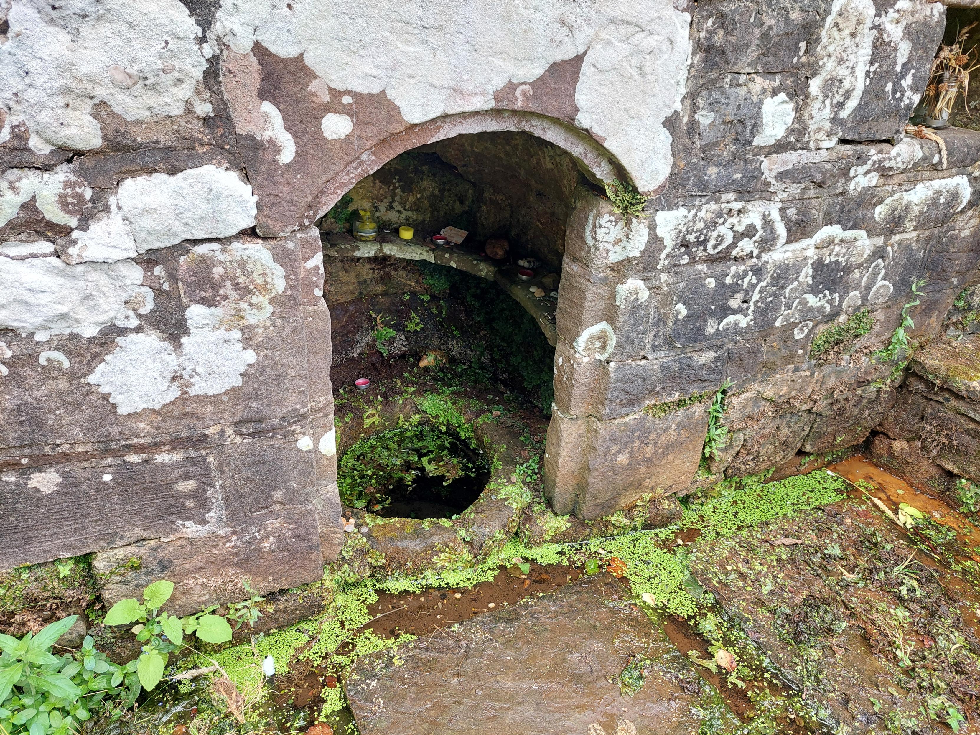 A small round Well sits inside an arched chamber in a lichen-encrusted stone wall. There are green water plants around the edge of the Well and between the flagstones in front of it. And, just visible on a tiny shelf inside the arched alcove, are candles and offerings from recent visitors