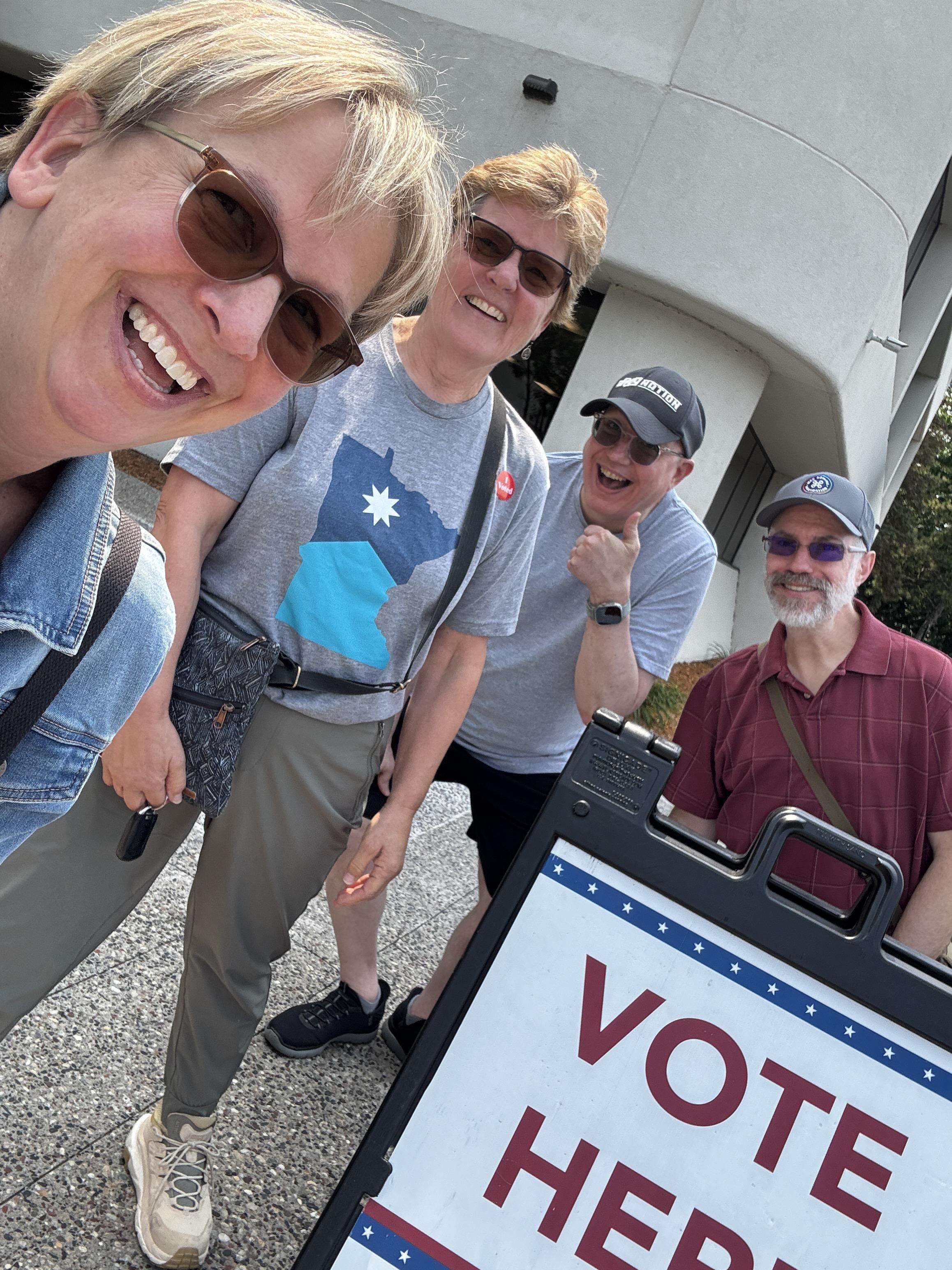 Four people standing near a Vote Here sign. Smiling like goobers.