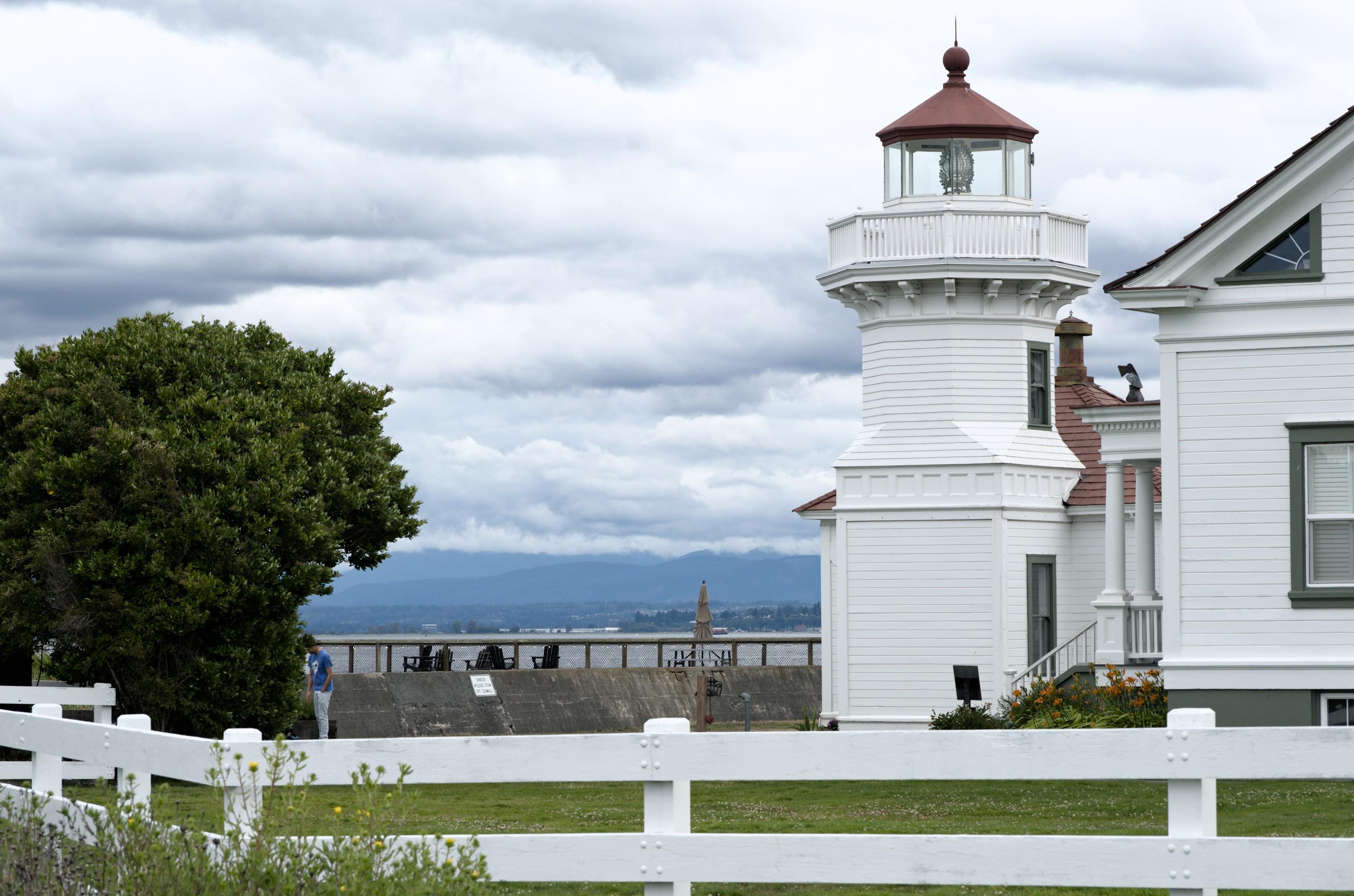 A short, stocky lighthouse with exterior made of painted white wooden boards, red tiled roof, stands on a green patch among similar looking houses, not really standing out. There's a white fence around them all. The heavy dramatic clouds fill the background.