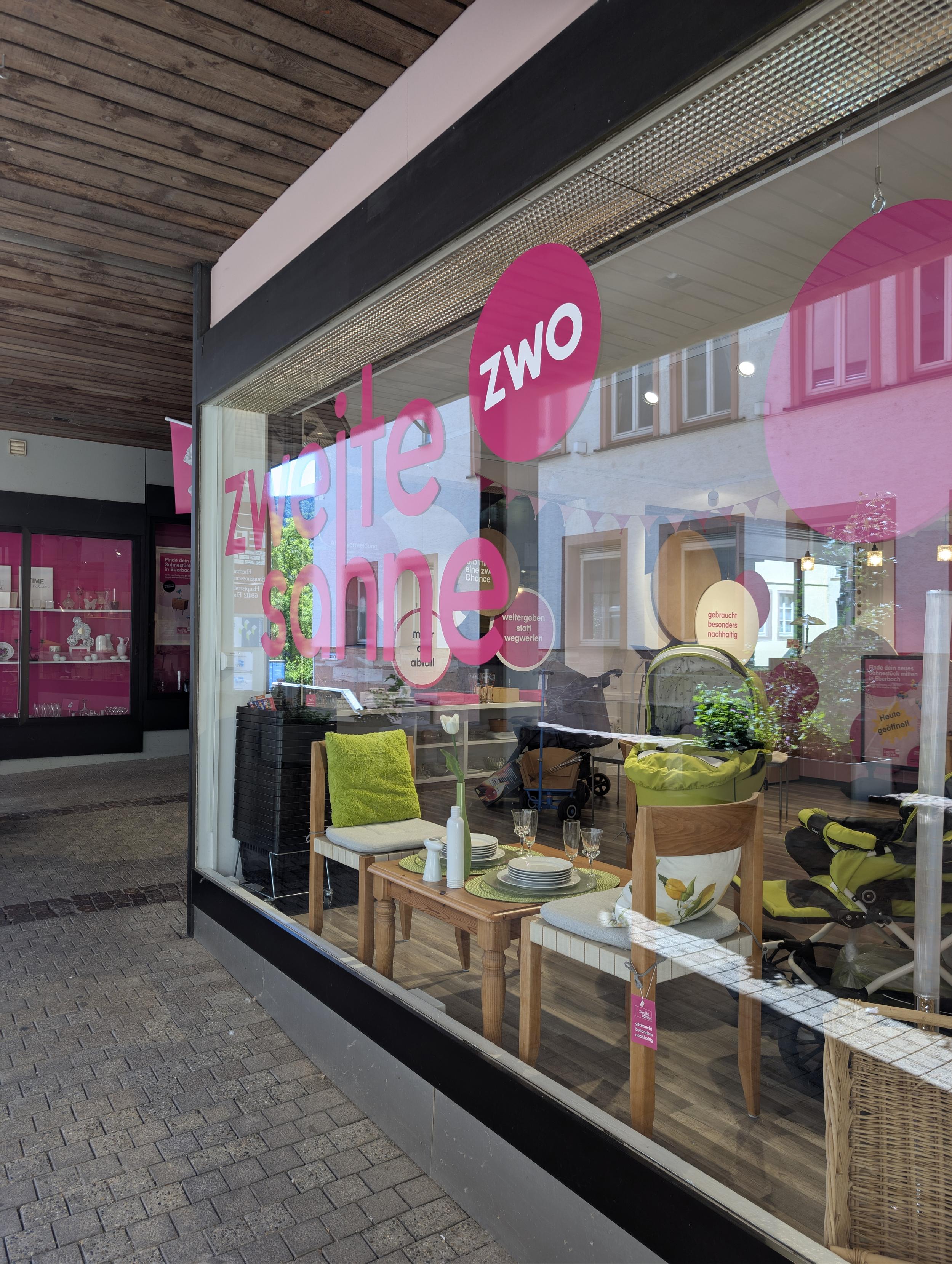 Das Schaufenster eines Geschäftes, in pink sind die Worte zweite Sahne aufgeklebt. Ausgestellt sind unter anderem zwei Stühle aus Holz mit Sitzflächen aus hellem Stoff, ein niedriger Holztisch mit hellem Geschirr und grünen Accessoires.
