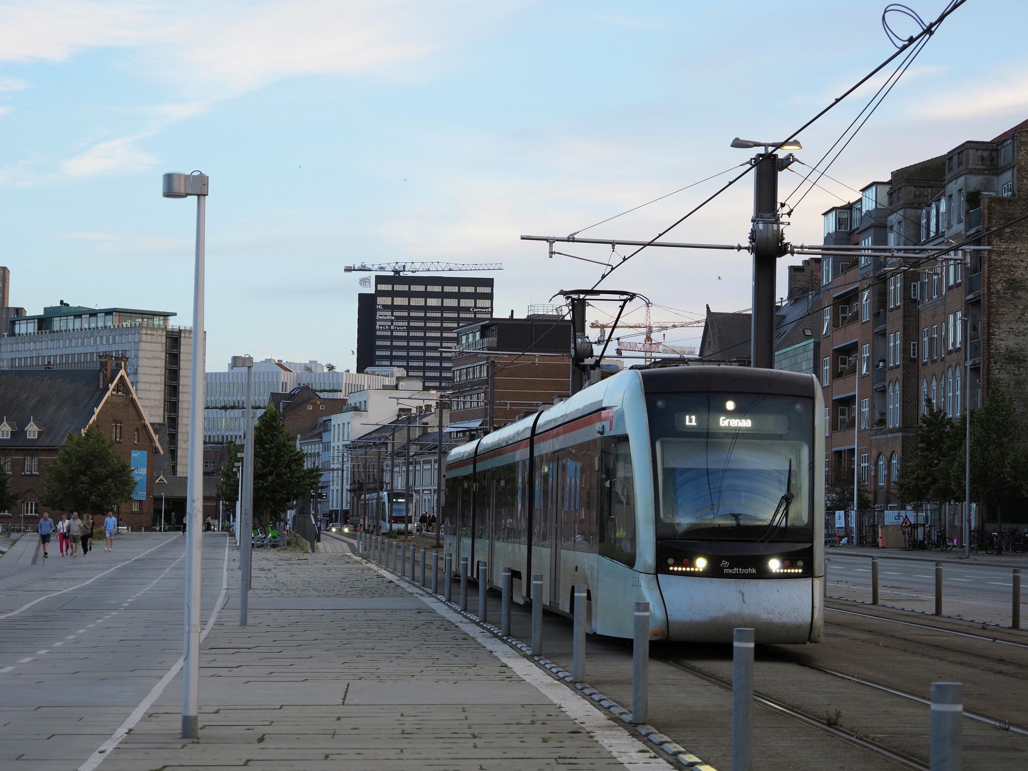 An oncoming Stadler Tango tram consisting of three articulated units, light-grey livery, destination indicator L1 to Grenaa, concrete double tracks, mixed modern and old industrial architecture in the background