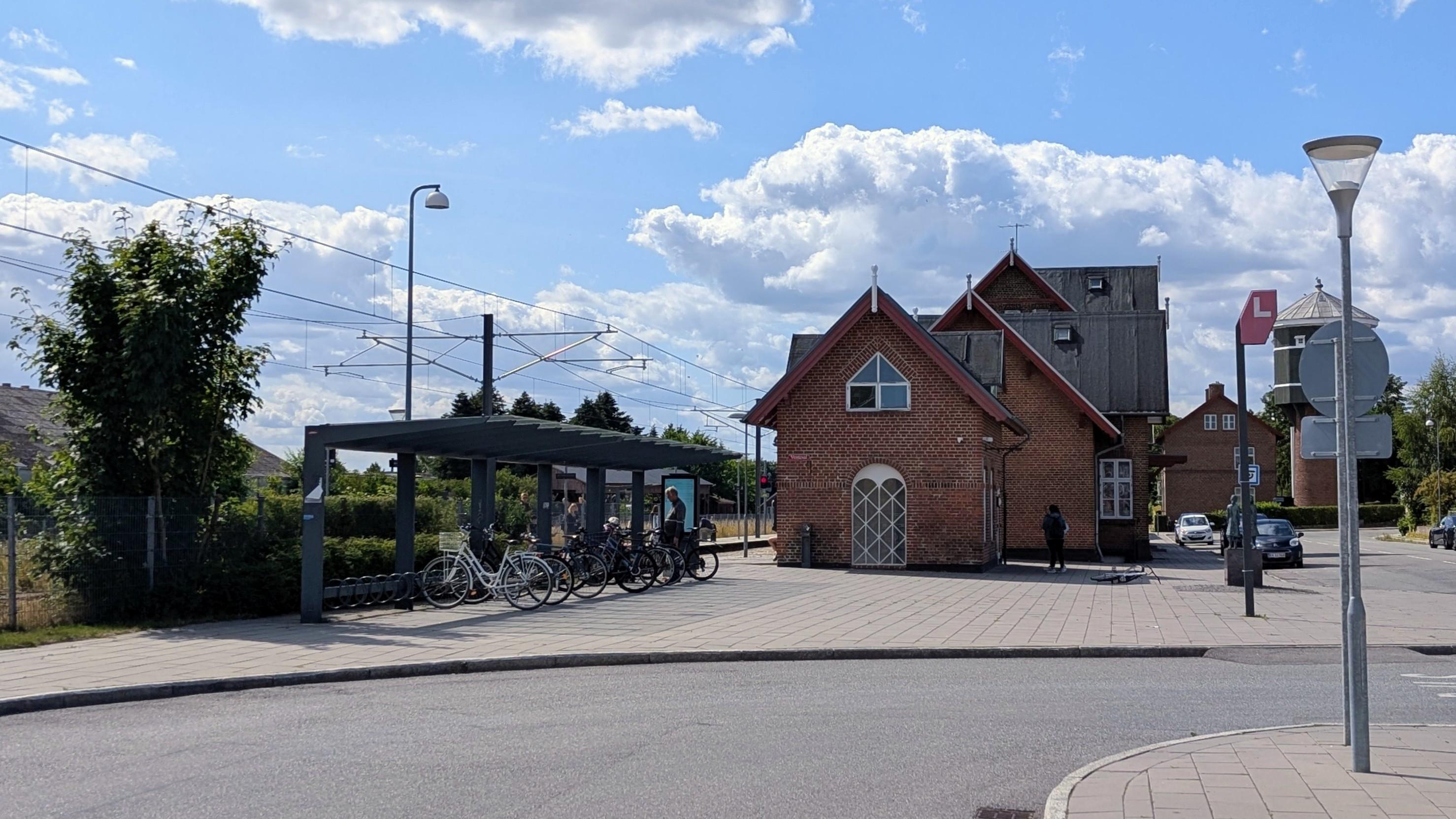 A brick station building with gabled roof, paved surroundings, bike parking, blue sky