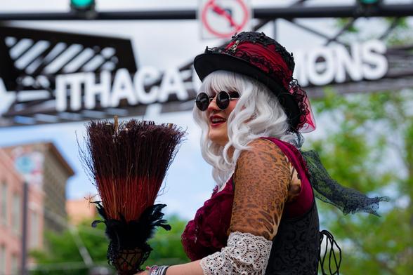 Woman with white hair and dark circular glasses and lacy clothes and hat holds up a broom under the ITHACA COMMONS sign