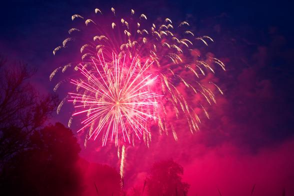 Pink firework radiates lines out from center behind a golden sparkly firework that fired first with clouds illuminated pink