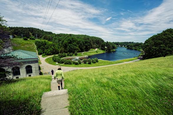 Looking down a grassy green slope with a small stripe of concrete stairs with a person in green walking down and past that the beginning of a river on the right and some kind of big square building to the left