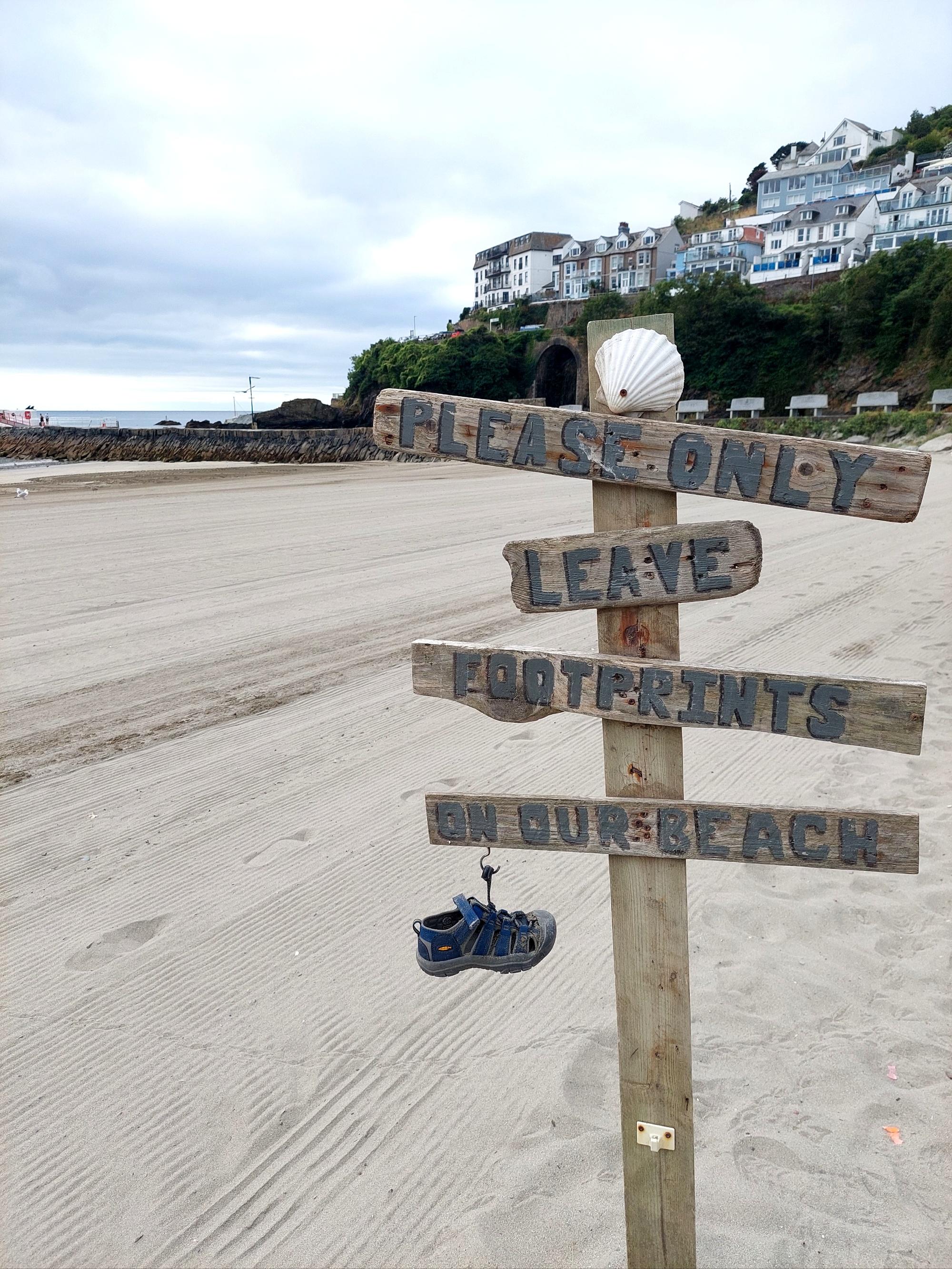 Set against a sandy beach with a harbour wall, headland, and houses in the background, a driftwood sign reads 'Please only leave footprints on our beach'. A child's shoe hangs from a hook on the bottom of the sign