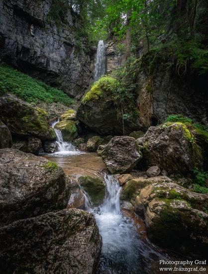 This image captures a serene and enchanting scene of a waterfall nestled in a lush, forested canyon. The waterfall cascades gracefully over moss-covered rocks, its clear water creating a sense of movement and tranquility. The falls are surrounded by rugged rock formations, some of which are covered in vibrant green moss and patches of grass, adding texture and color to the scene.
The water flows down in multiple streams, creating a dynamic and picturesque composition as it splashes into a shall…