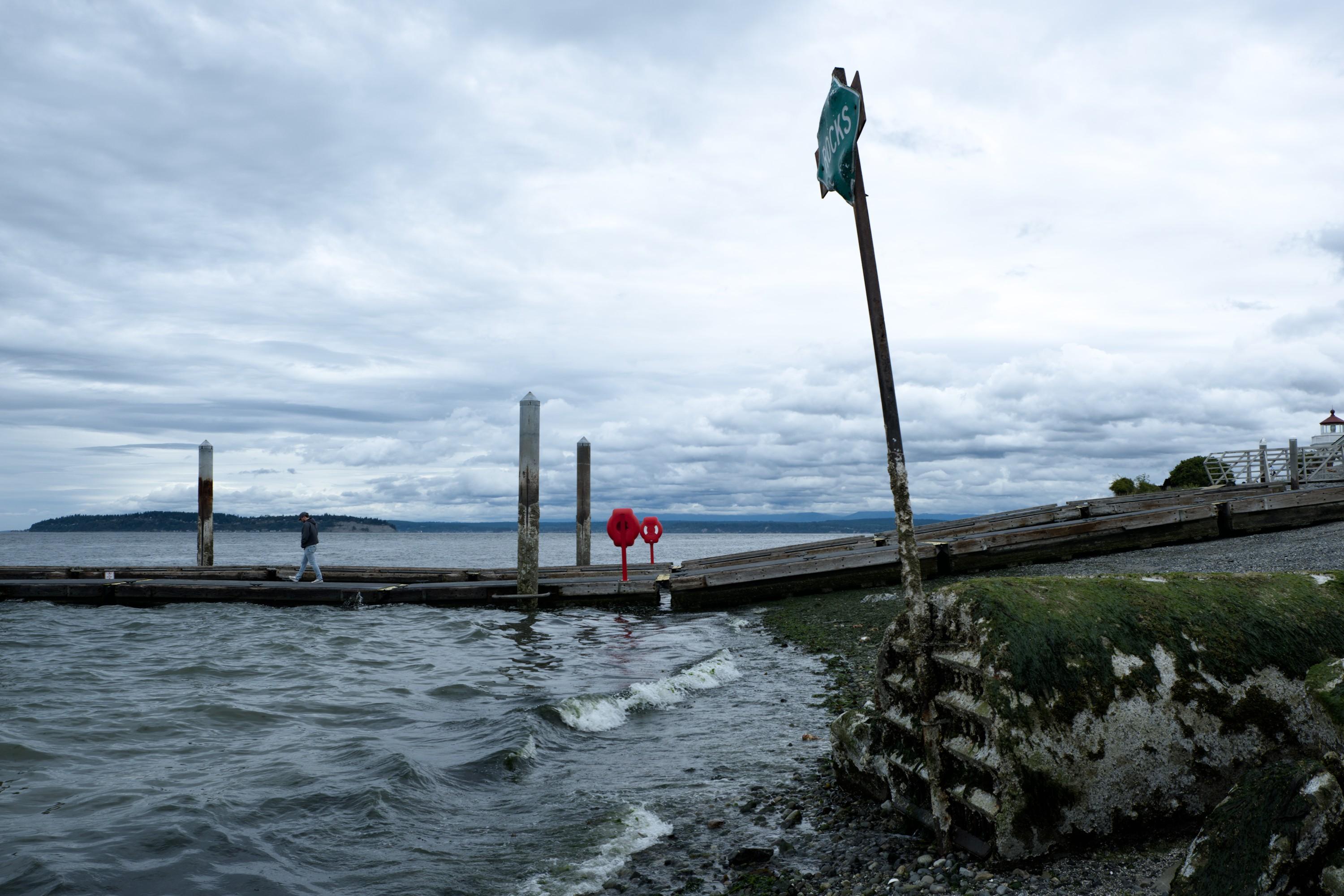 Windswept rocky beach with an old stony pipe covered in moss, a tilted rusty sign saying 'Rocks' and a small dock with two bright red life saver stands. The sky with heavy, gray, dramatic clouds oppresively dominates the photo.
