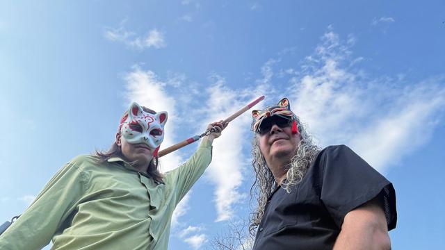 On the left a person in a light green shirt holds up two sections of a three section staff,  he's wearing a light colored fox mask,  the guy on the right has a black shirt and would look like a professional wrestler if we here larger and more muscular -- above them a blue sky with high altitude fair weather clouds