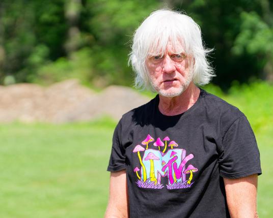 Rather annoyed looking older man with all white hair and a black mtv shirt suggestive of psychedelic mushrooms with a blurred out grass field, maybe gravel piles and trees in the far background