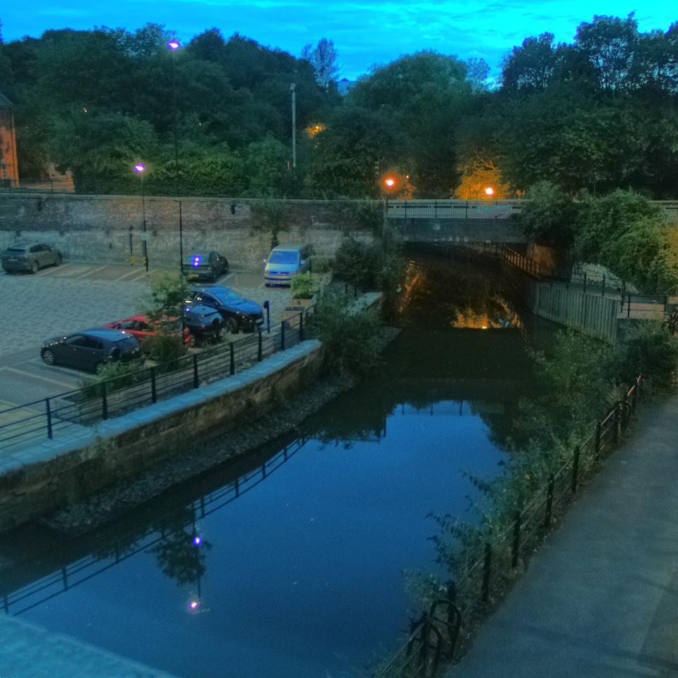 Photograph of the tidal stretch of Ouseburn River at south side of Ouseburn Bridge in Newcastle upon Tyne. To the left (south-west) of the bridge is Cut Bank, and to the right (north-east) is Byker Bank. In the foreground is the pedestrian Riverside Walk and the photo also spans some surrounding built-up areas and trees, with sky in the background. Still image captured from webcam at 04:44 hrs on Thursday 30 July 2026.