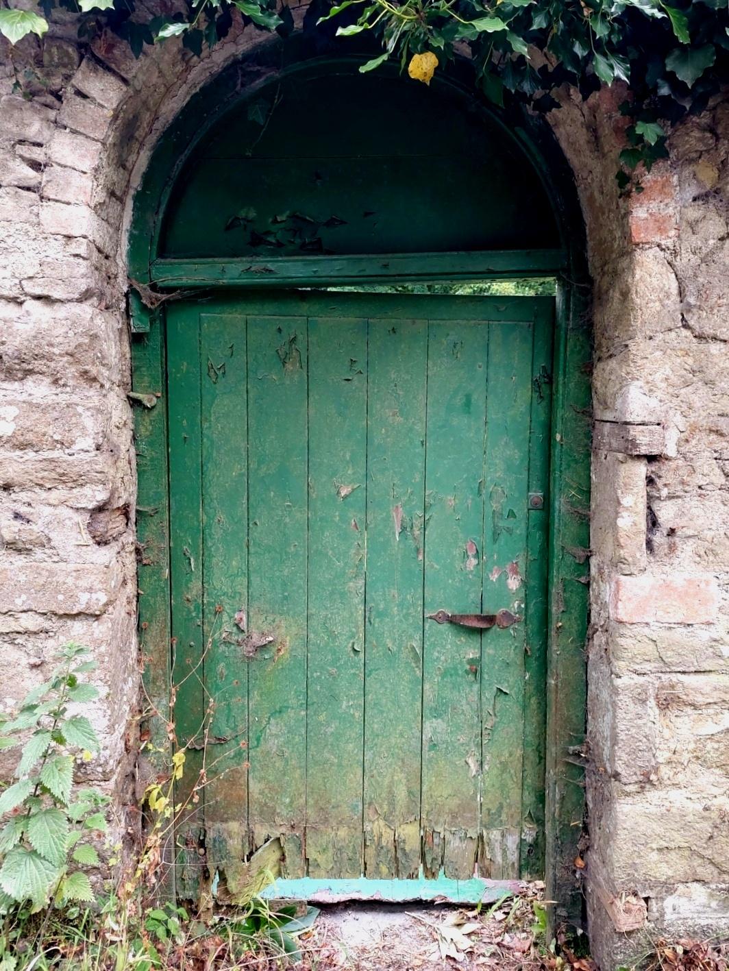 An old and weathered green door hangs at a slight angle in a stone wall archway