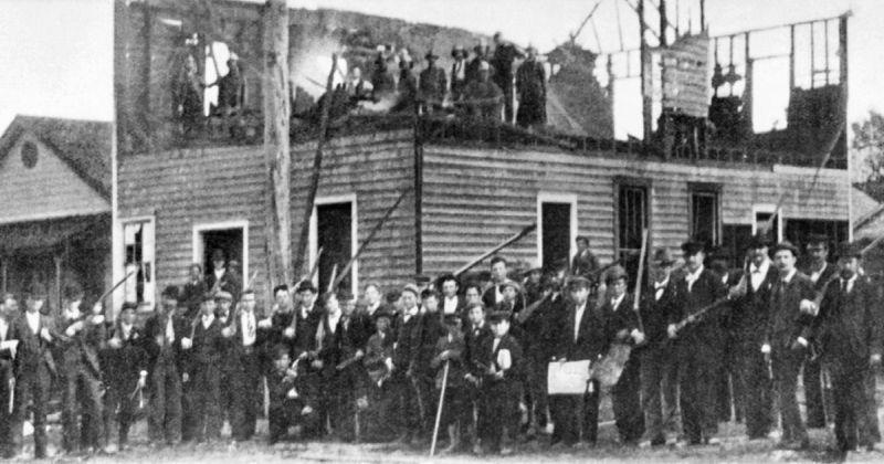 In the Wilmington Massacre of 1898, dozens of men stand in front of and on top of a burned-out building. They are dressed in dark suits, coats, and hats, which were common in the late 19th century. Many of them hold long rifles or shotguns. This white supremacist mob is celebrating after destroying The Daily Record, the only Black-owned and edited newspaper in the city.