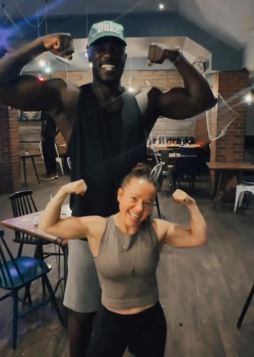 the actors in the scene without their costumes flexing their biceps in the gym, a small woman and a large man
