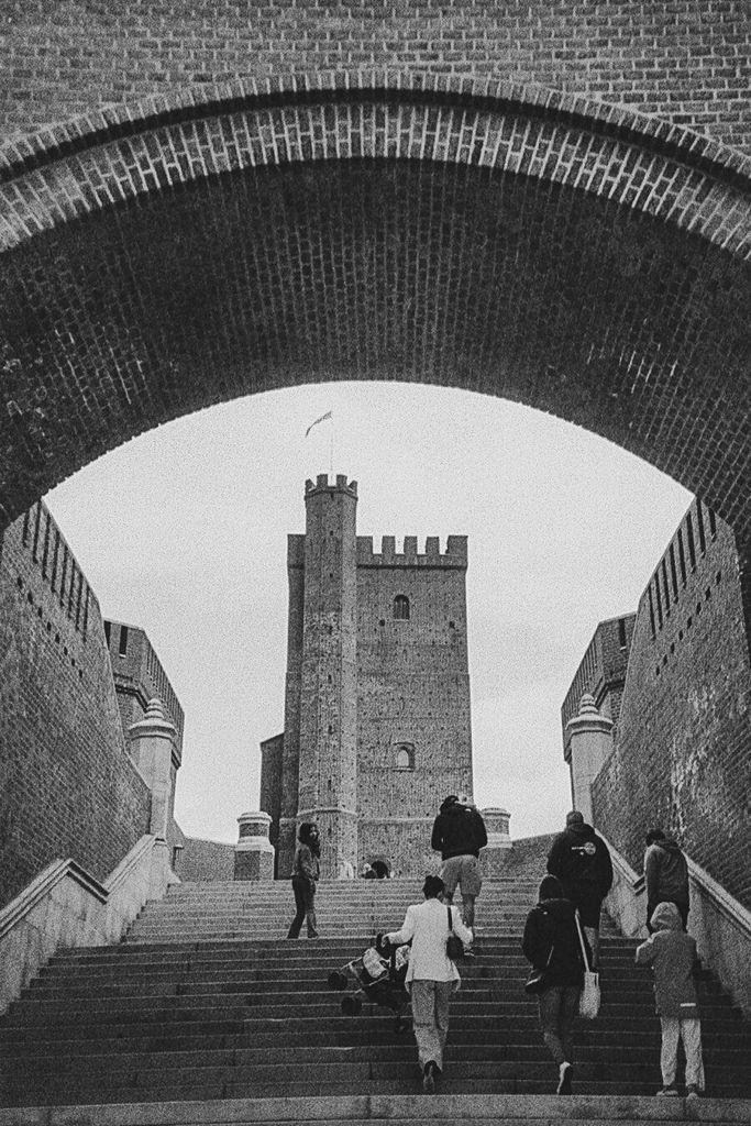 Visitors climb wide stone steps beneath a massive brick arch toward a tall crenellated medieval tower with a small flag, captured in high-contrast black-and-white with visible film grain.