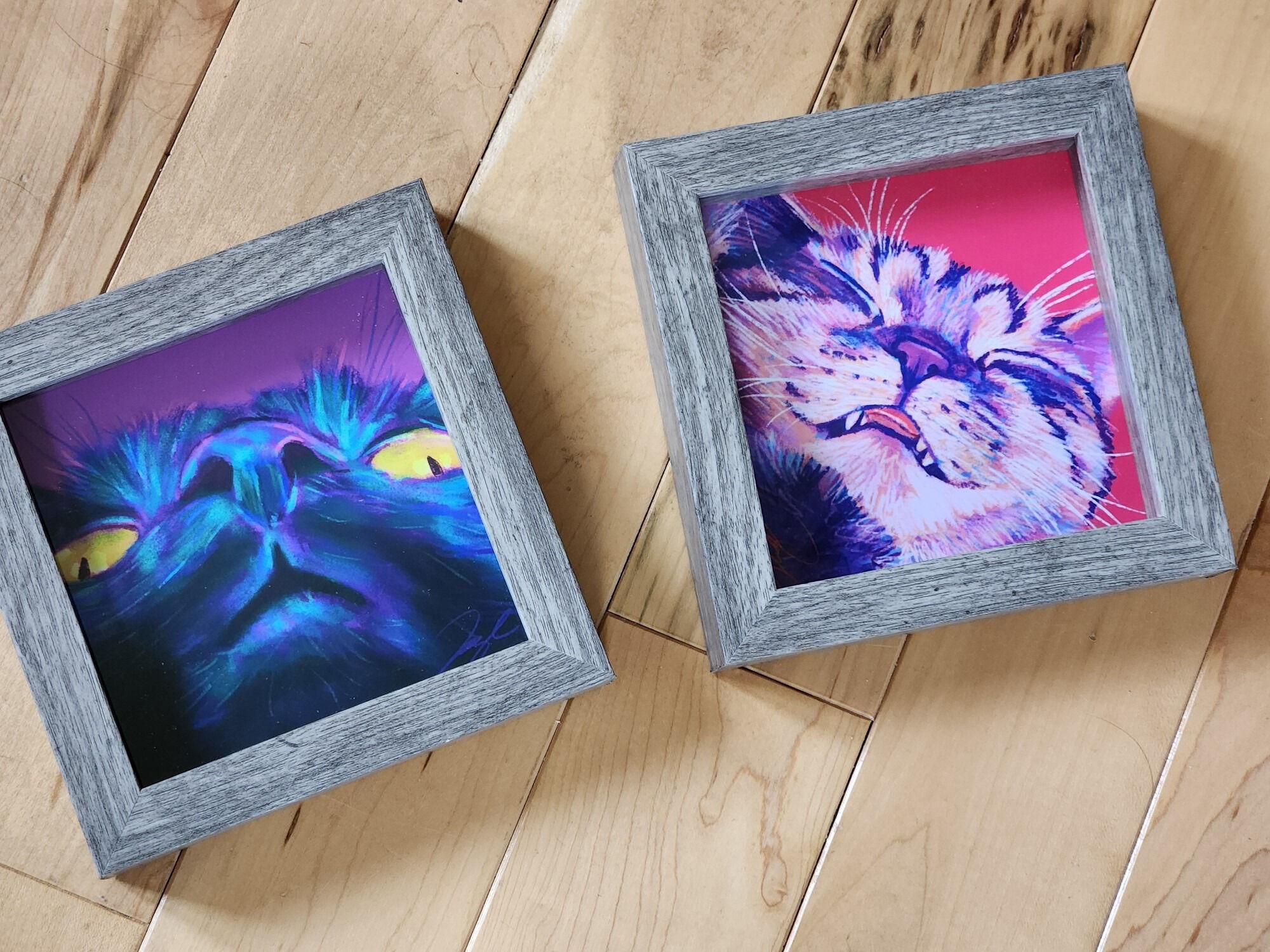 Photo of two framed paintings of cats painted by JoyousJoyfulJoyness. The left is a black cat with golden eyes and the right is a sleepy cat. Source:  @culturalgutter.bsky.social
