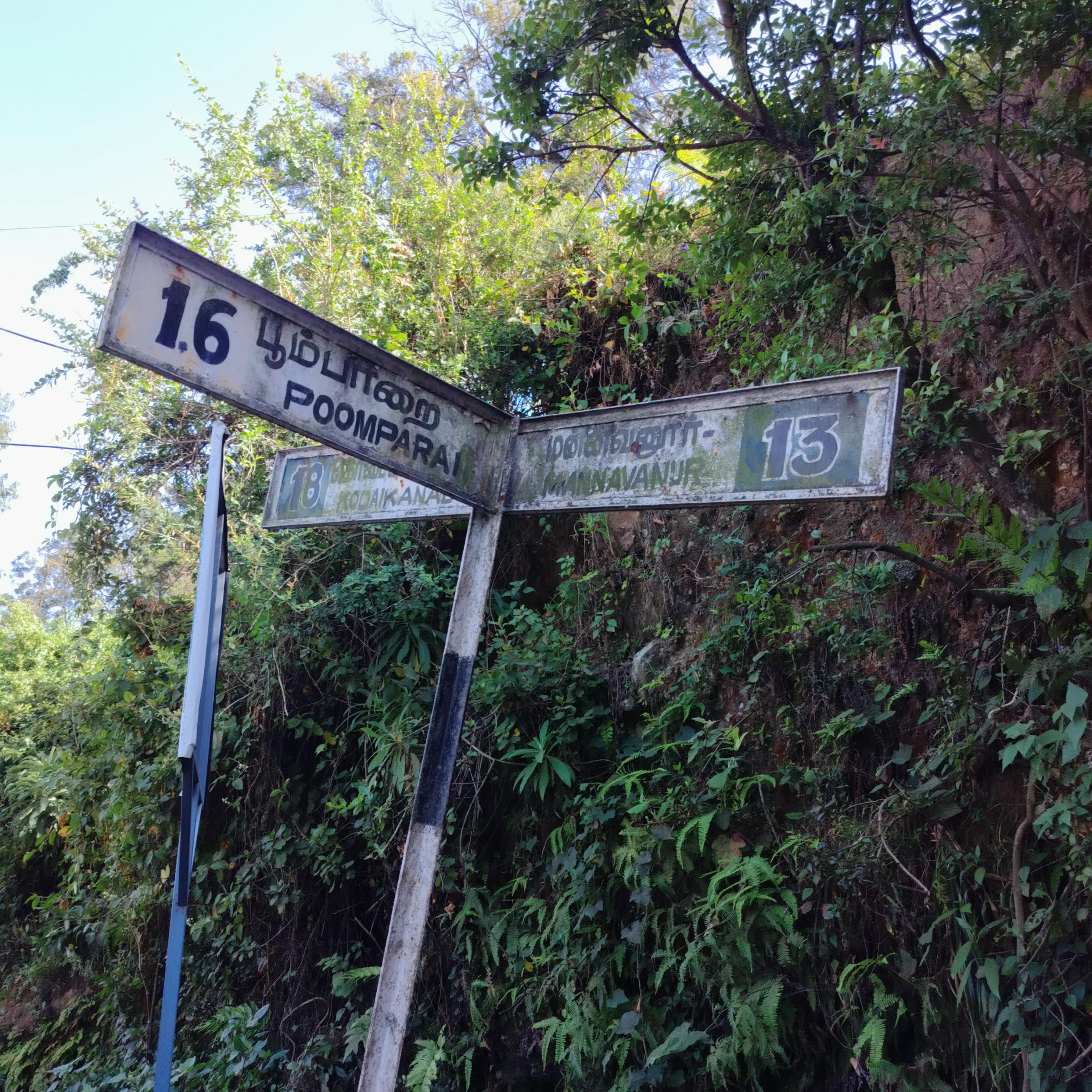 A weathered metal road sign at a junction, surrounded by lush green foliage. The sign indicates directions to Poomparai (1.6 km) and Mannavanur (13 km), with a partially visible sign pointing towards Kodaikanal (18 km).