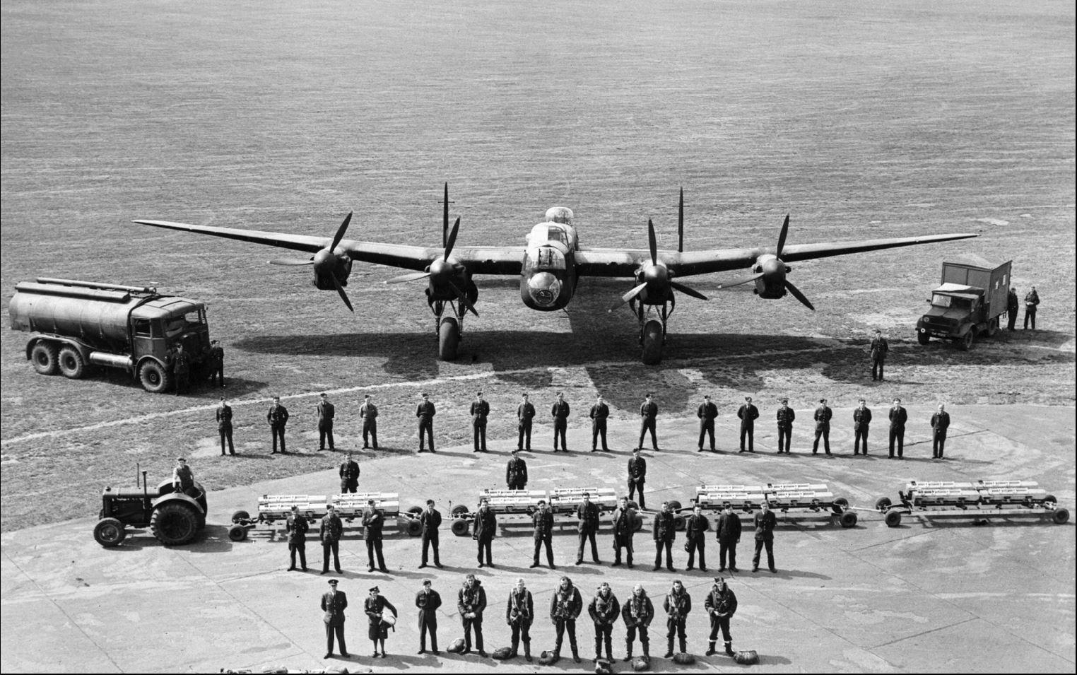 View from high up (perhaps from a control tower) of the flight crew and ground crew for an Avro Lanacater bomber, lined up in front of it. There are 48 personnel shown, of whom 7 appear to be flight crew. Aside from the bomber, there are 3 other vehicles: a fuel truck, a tractor pulling a train of 4 bomb carriages, and another service truck.