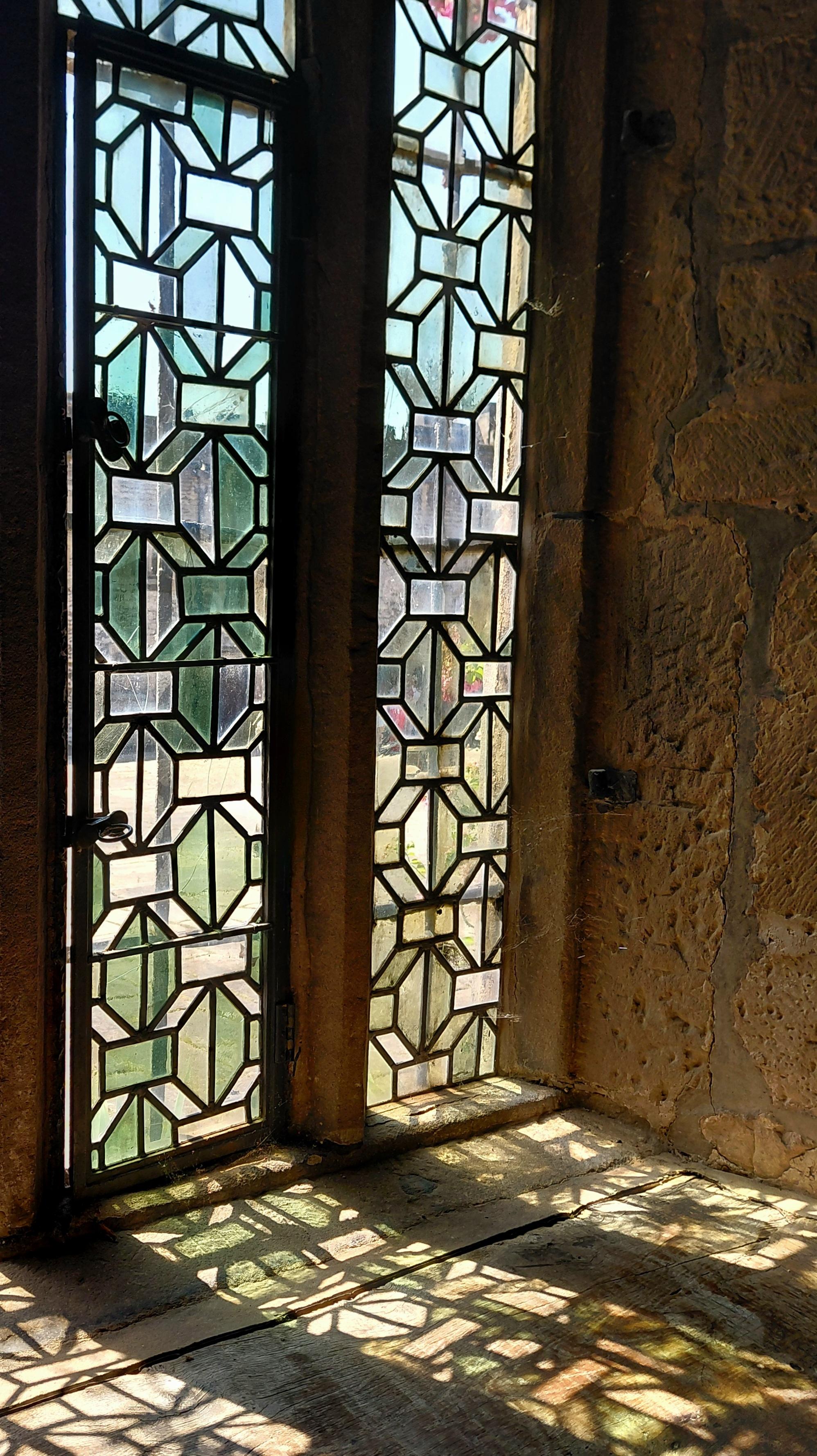 Close up of a stained glass window in a medieval / tudor parlour. The window features a lattice work pattern of opaque and transparent panes of glass in green and white. Light shines through the window from the castle courtyard outside, spilling coloured squares onto the wide wooden window sill.