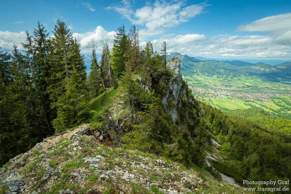 This image captures a breathtaking and expansive mountain landscape, showcasing the serene beauty of a high-altitude vista. The foreground features a rugged, rocky cliff adorned with patches of green vegetation, including moss and small shrubs, adding texture and a sense of wildness to the scene.
Beyond the cliff, the landscape opens up to reveal a vast expanse of dense forests and rolling hills. The forests are dominated by tall evergreen trees, their dark green foliage creating a rich and vib…