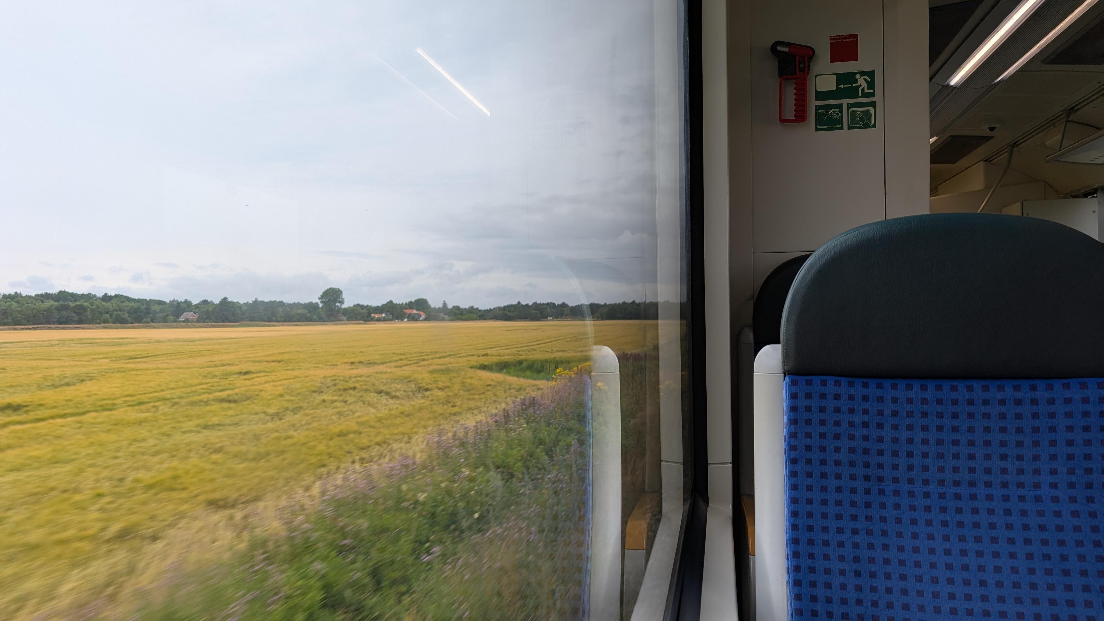 Train scene, seat with blue padding, big window, a field, trees and a house in the background