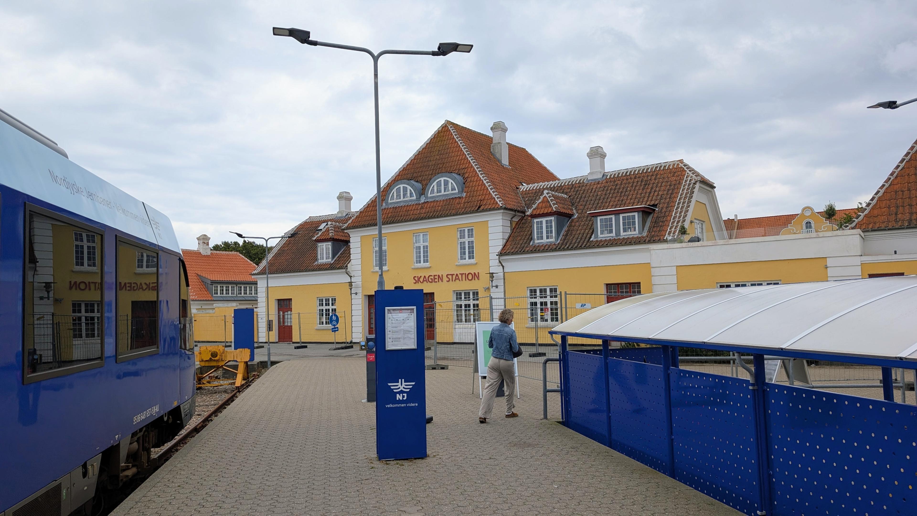 A yellow station building with white lattice windows, tiled platform, a blue NJ LINT 41 DMU
