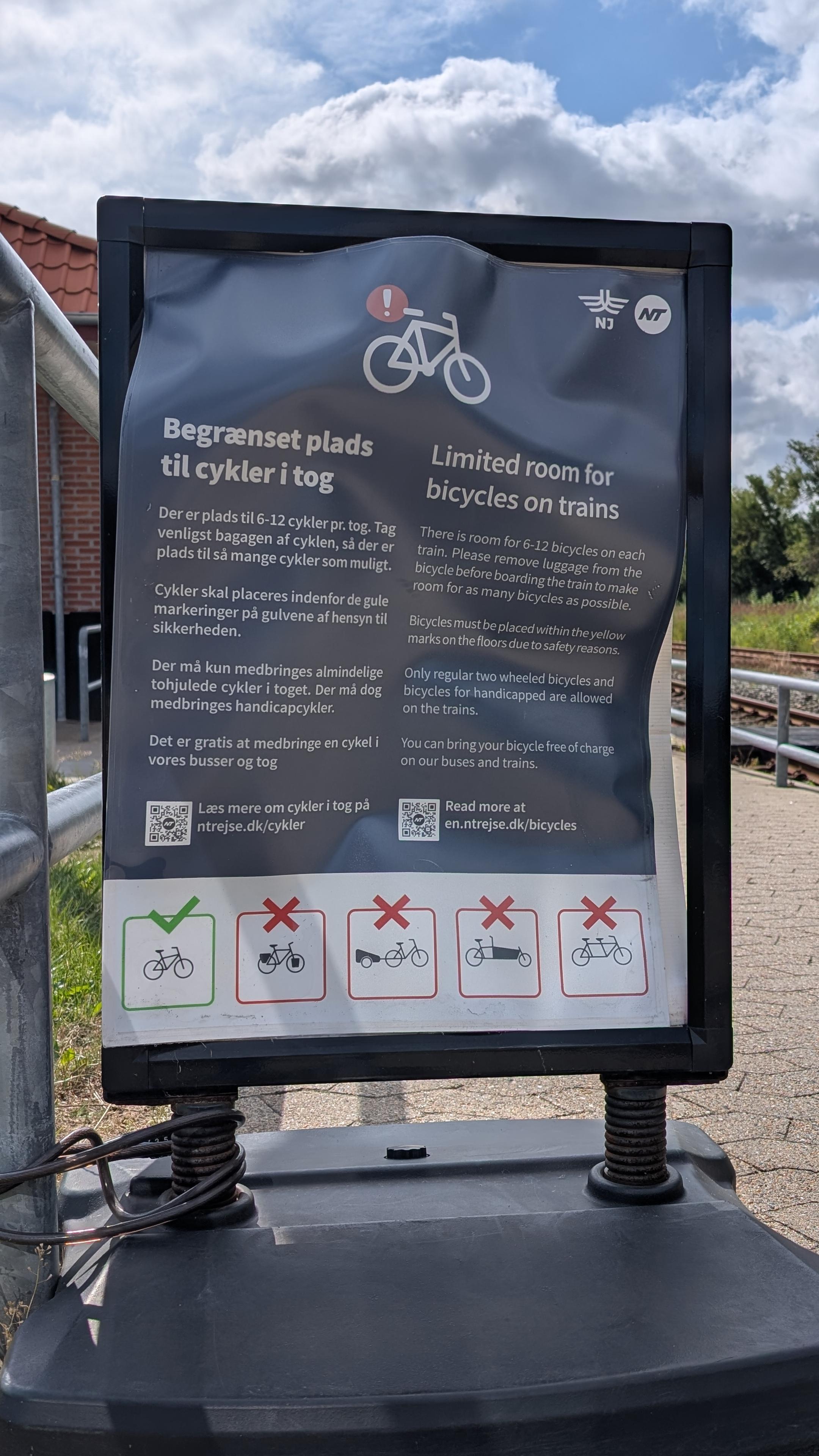 Stand-up sign: 'Begrænset plads til cykler i tog - Der er plads til 6-12 cykler pr. tog. Tag venligst bagagen af cyklen, så der er plads til så mange cykler som muligt. Cykler skal placeres indenfor de gule markeringer på gulvene af hensyn til sikkerheden. Der må kun medbringes almindelige tohjulede cykler i toget. Der må dog medbringes handicapcykler. Det er gratis at medbringe en cykel i vores busser og tog. Læs mere om cykler i tog på ntrejse.dk/cykler

Limited room for bicycles on trains - There is room for 6-12 bicycles on each bicycle before boarding the train to make train. Please remove luggage from the room for as many bicycles as possible. Bicycles must be placed within the yellow marks on the floors due to safety reasons. Only regular two wheeled bicycles and bicycles for handicapped are allowed on the trains. You can bring your bicycle free of charge on our buses and trains. Read more at en.ntrejse.dk/bicycles'