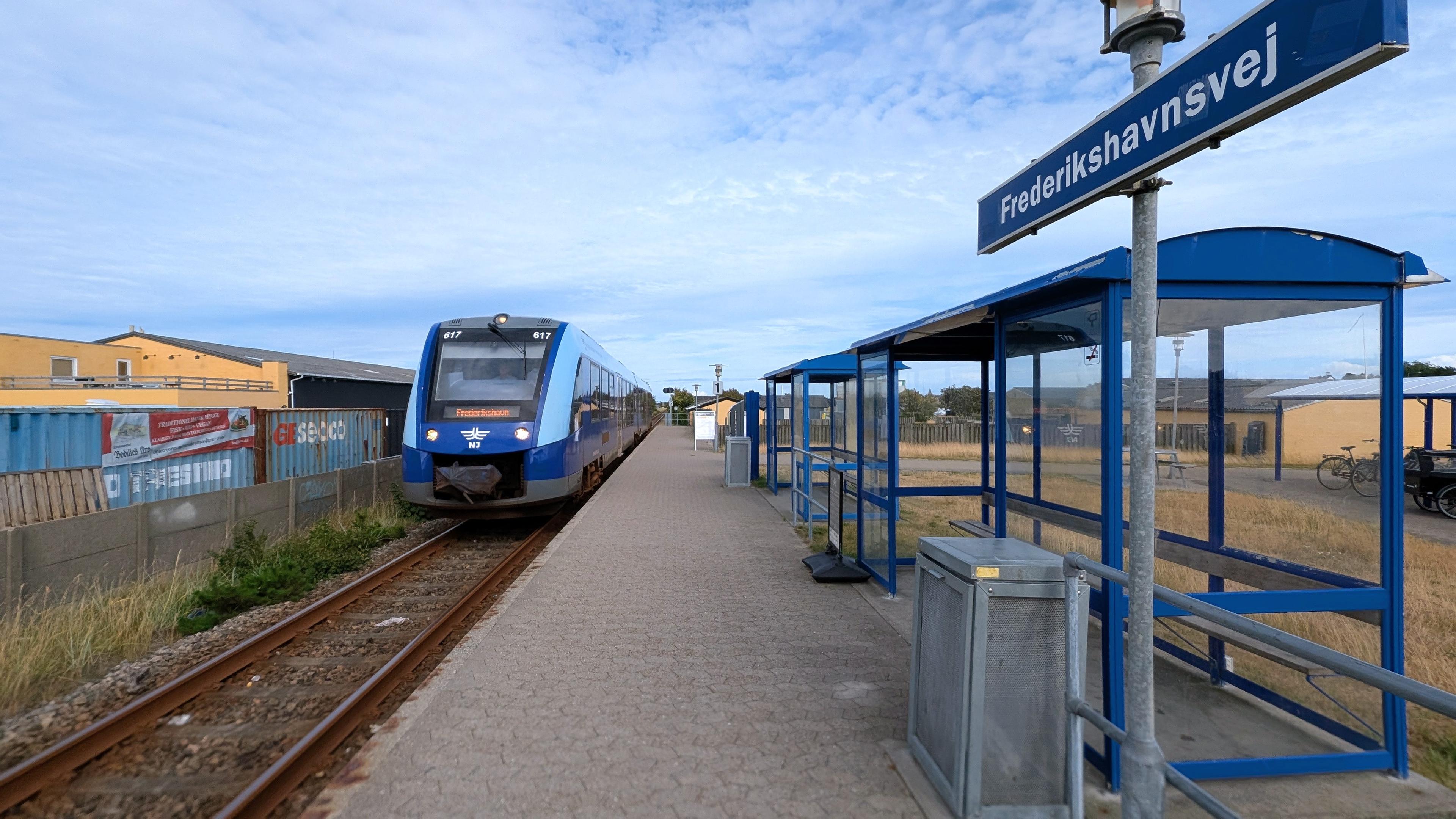 A train stop with a tildmed platform and a small shelter, sign "Frederikshavnsvej", arriving NJ LINT 41 DMU at platform