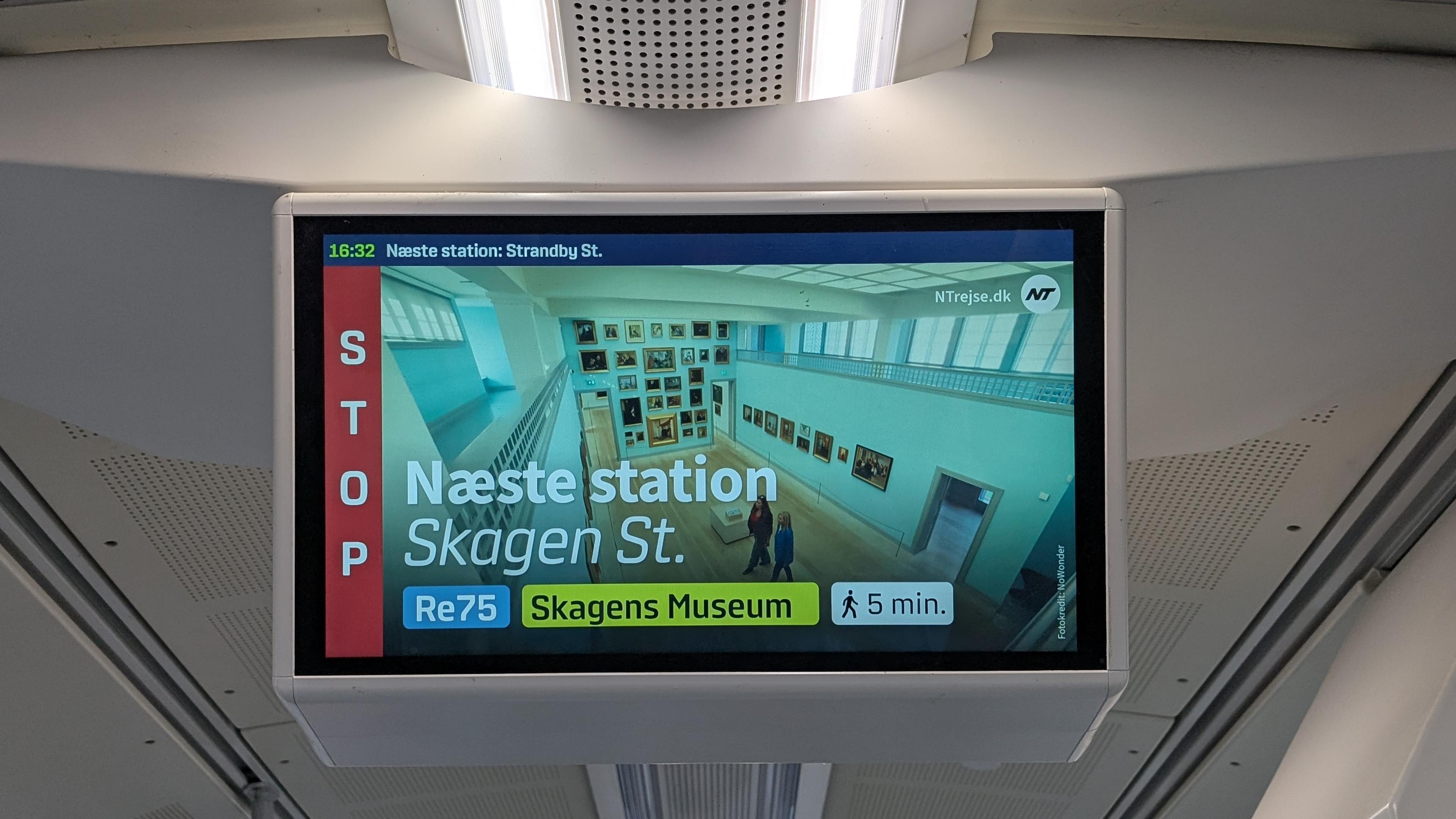 A passenger information screen: Tiny row on the top '16:32 Næste station Strandby St.', a red column on the left underneath 'Stop', most of the space filled with a picture of an art museum hall 'Næste station: Skagen St.' as well as labels 'Re 75', 'Skagens Museum' and '5 min. walking distance'