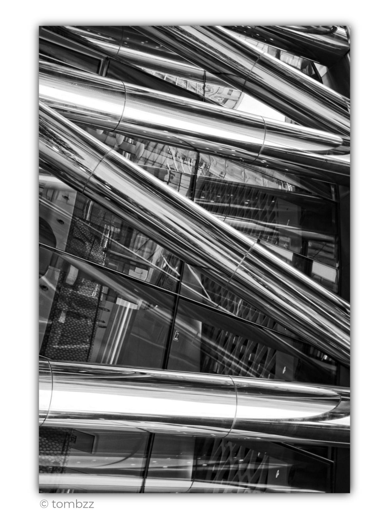 Black-and-white photograph of a fragment of an ultramodern façade made of glass and polished steel, with surfaces resembling chrome. The frame is filled with massive cylindrical structural elements running diagonally and intersecting at different angles. Their mirror-like surfaces reflect light, fragments of neighbouring architecture, geometric window divisions and additional metal beams, making the space appear layered and difficult to read at first glance.
Between the steel tubes, glass panels, dark frames, reflections of the building’s interior and small construction details are visible. The composition forms a dense network of gleaming lines, reflections and distortions, with almost no calm or empty areas. The strong contrast between bright highlights on the metal and the darker glass emphasizes the cool, industrial character of the architecture.
The image has been rotated by 90 degrees, creating a deliberate visual trick. Structural elements that may originally run vertically or horizontally now appear as dynamic diagonal beams. This shift disturbs the sense of gravity and orientation, transforming the façade into an abstract composition built from chrome-like tubes, glass, light and reflections.