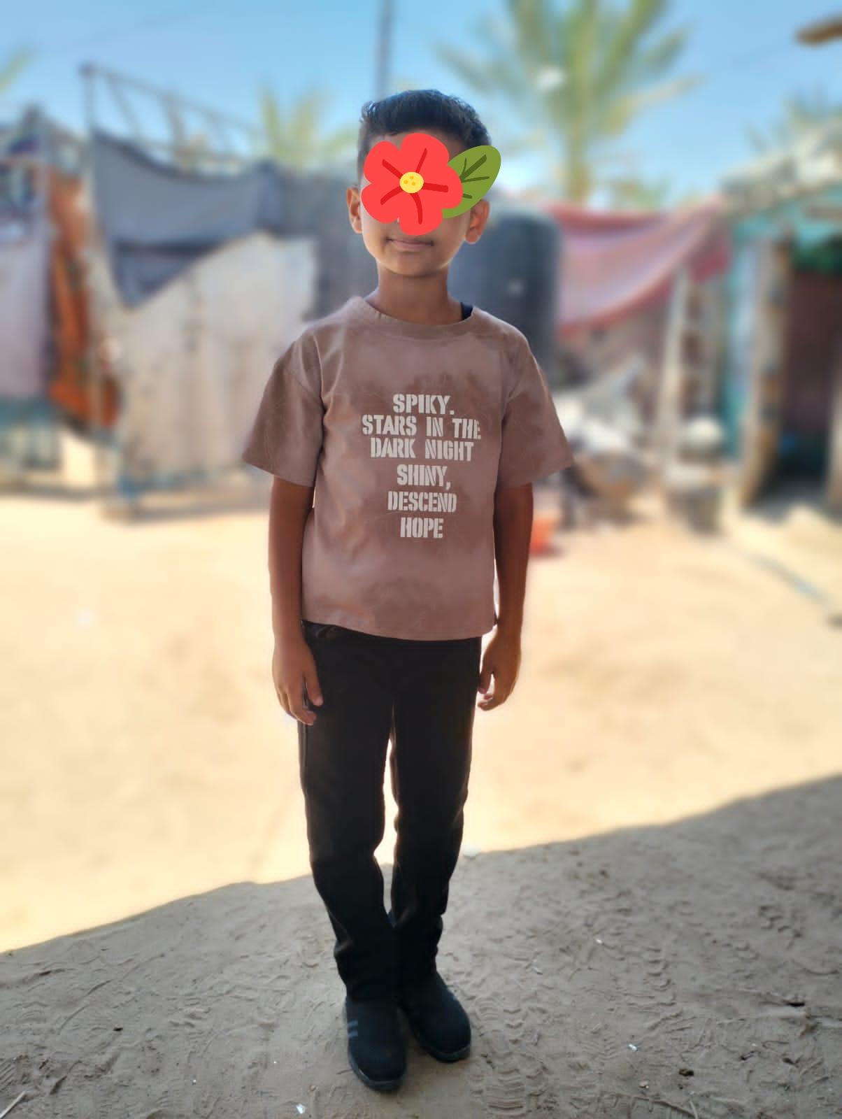 A child stands in front of blurred structures wearing a light brown t-shirt with the text "SPIKY. STARS IN THE DARK NIGHT SHINY, DESCEND HOPE" and black pants.