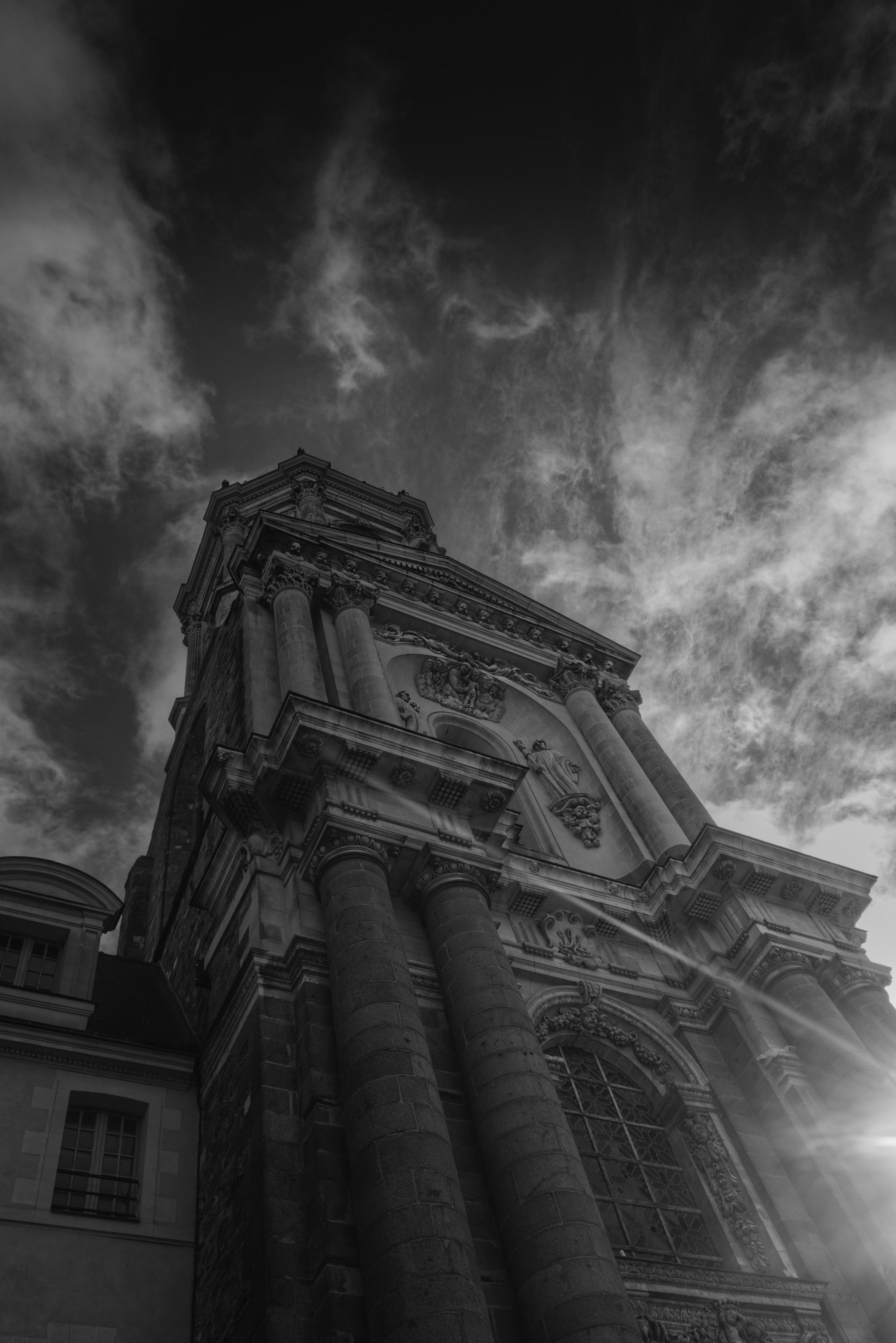 Photo en noir et blanc de la façade de l'église Saint-Melaine de Rennes, prise en contre plongée devant le ciel nuageux