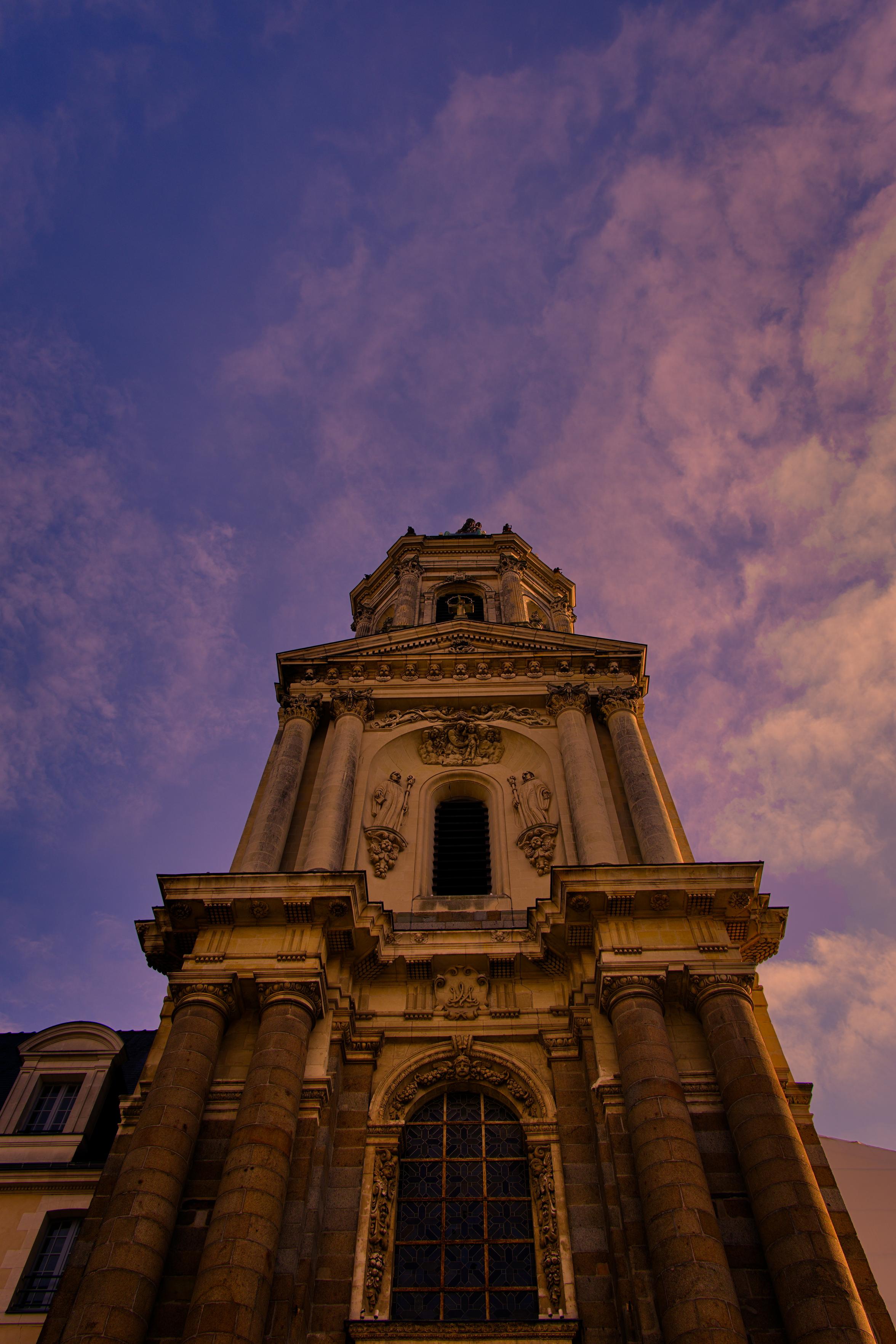 Photo en couleurs de la façade de l'église Saint-Melaine de Rennes, prise en contre plongée, la pierre apparaît ocre tandis que le ciel que le ciel bleu est parcouru de nuages roses