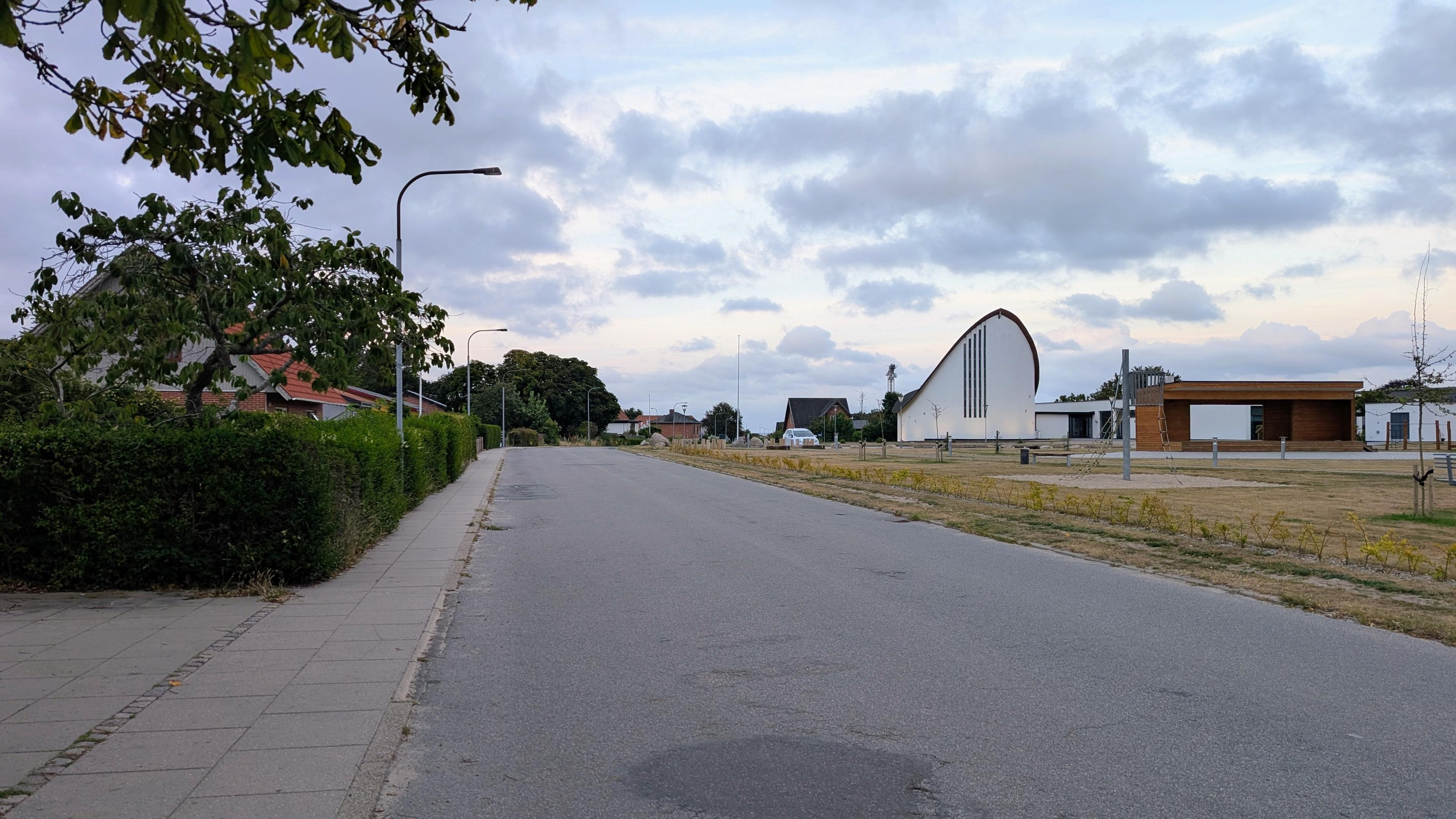 A wide street, a meadow with a modern church, houses on the other side of the street with hedges