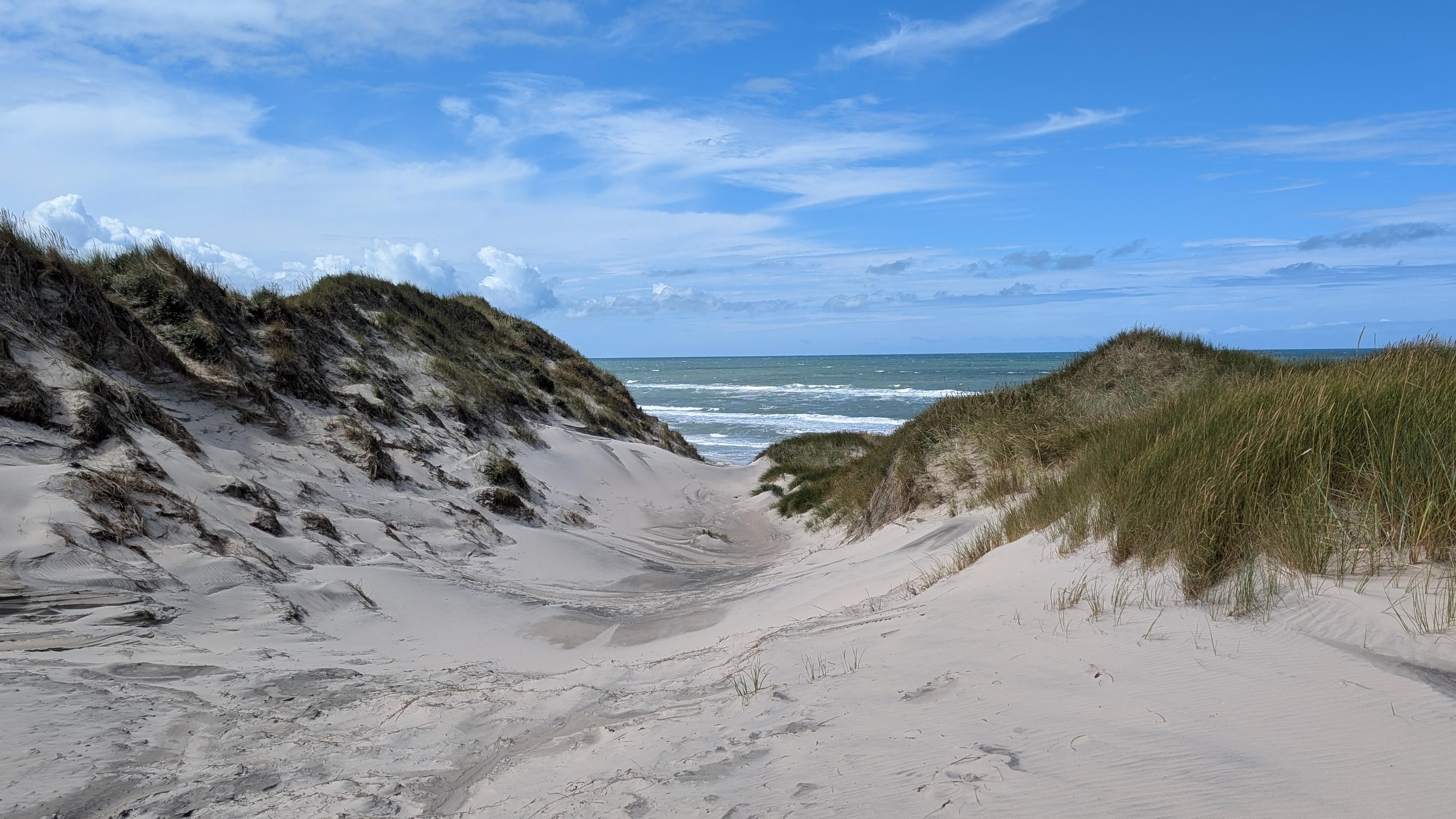 Sand dunes with grass covered tops, part of the sea visible through a 'trench', blue sky