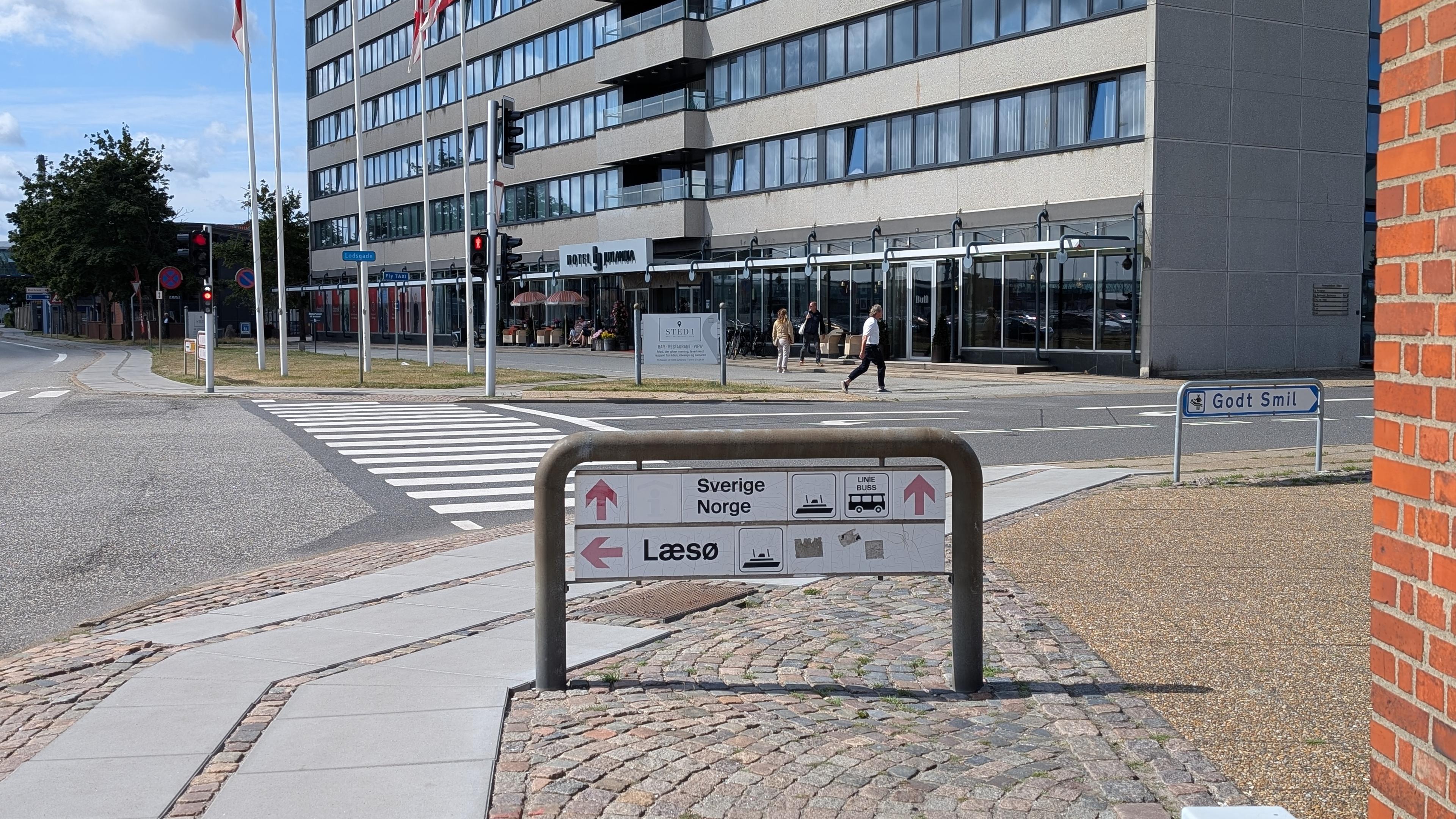 A white sign mounted with a metal frame to onto the pavement, black lettering and symbols, red arrows, Læsø ferry to the left, Sverige/Norge ferry and bus lines straight