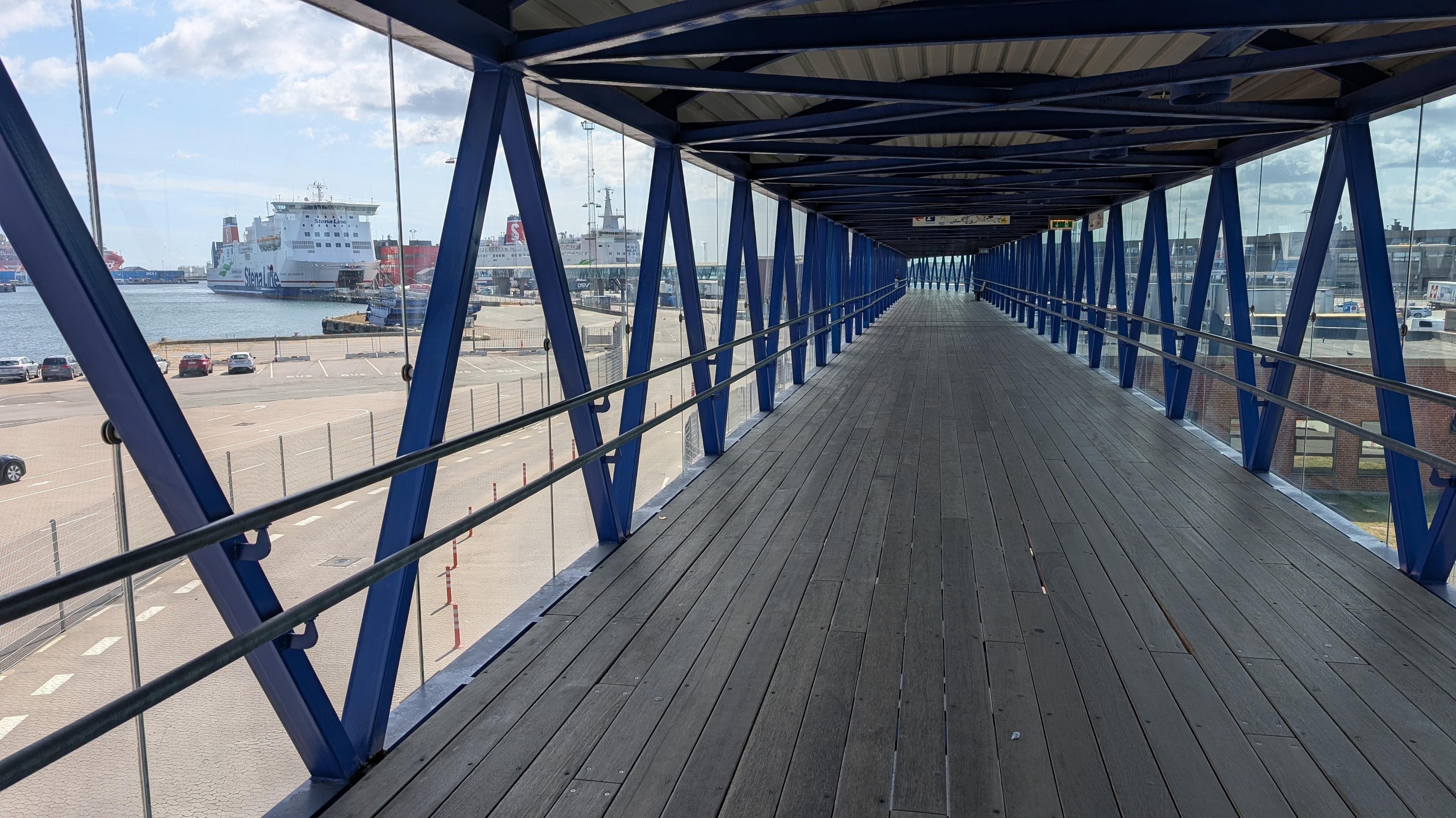 A passenger bridge seen from inside, blue diagonal metal bars along glas panes on both sides, wooden floor, Stena ferries can be seen outside