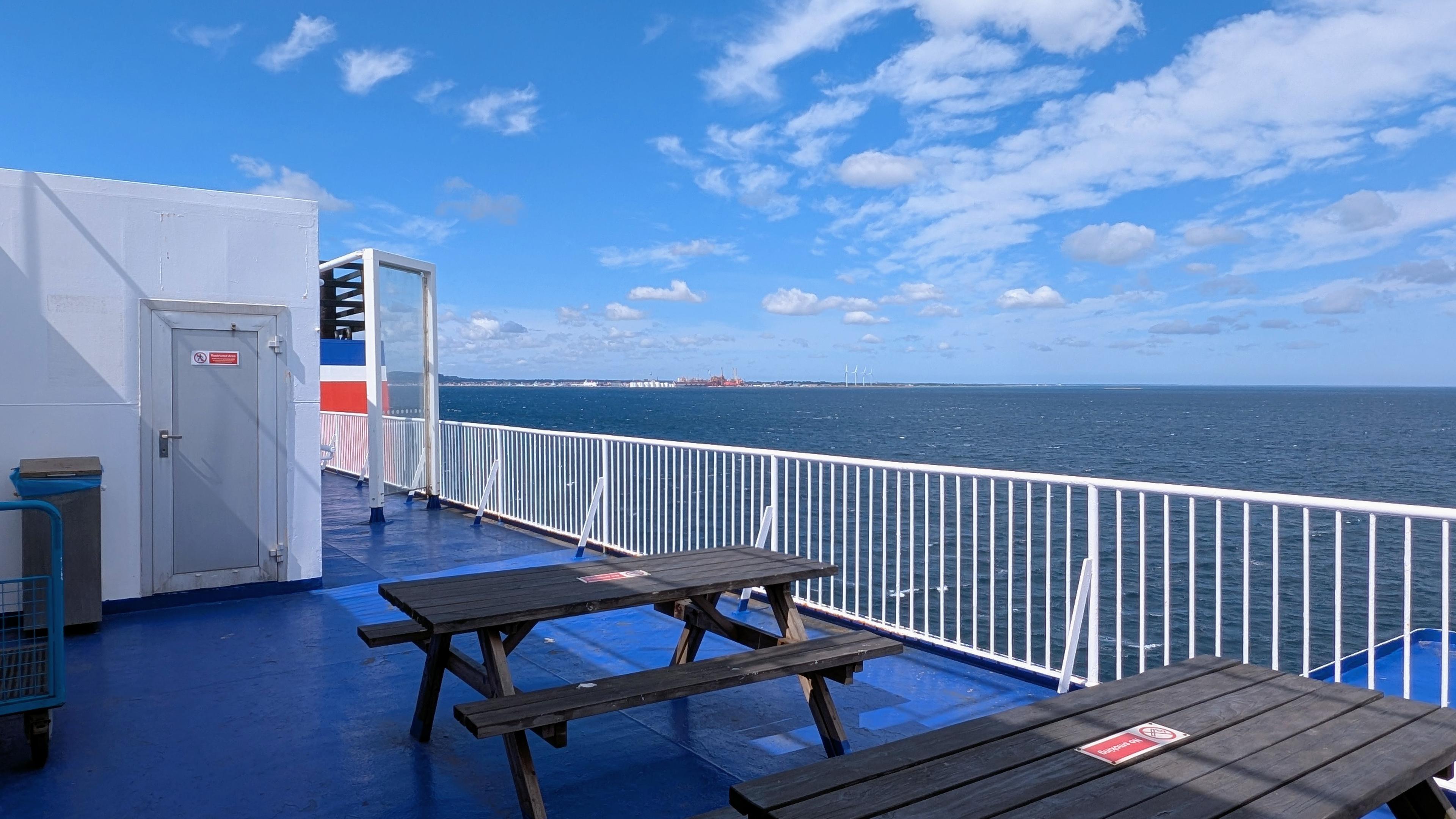 Wooden benches and tables on an outside deck, blue floor, white railing, blue sea and sky, some clouds