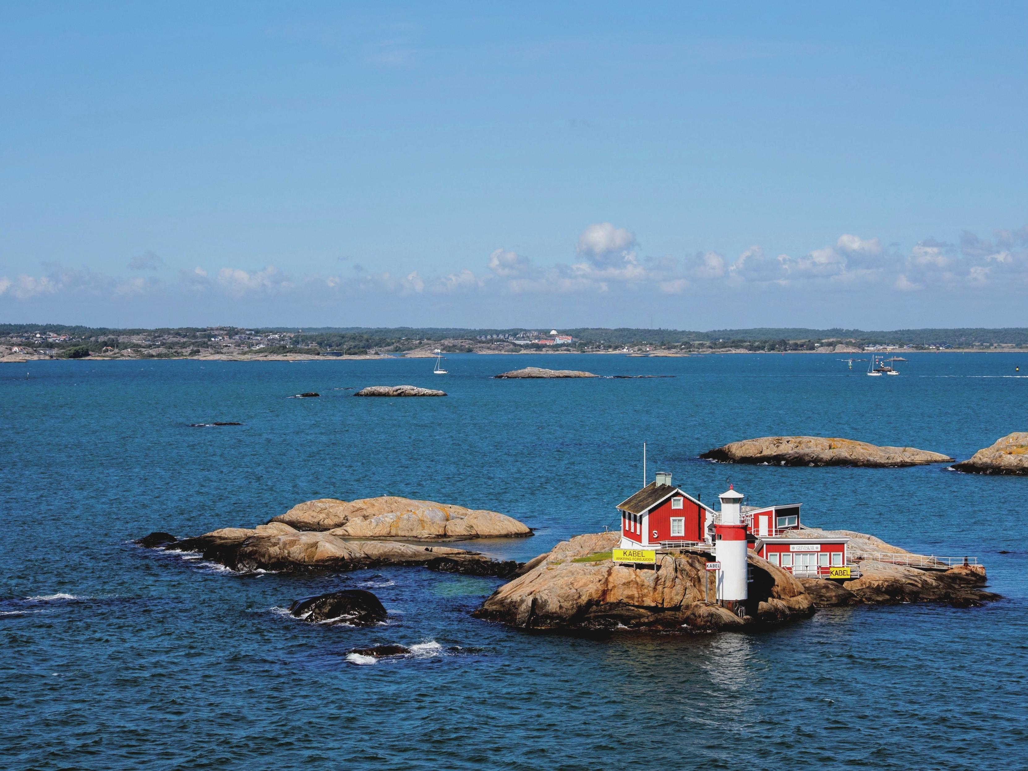 Rather smooth, polished rocks peaking out of the blue sea, small red-white houses and a small light house on one of the rocks in front, in the background some white boats and a forested flat landscape with patches of buildings