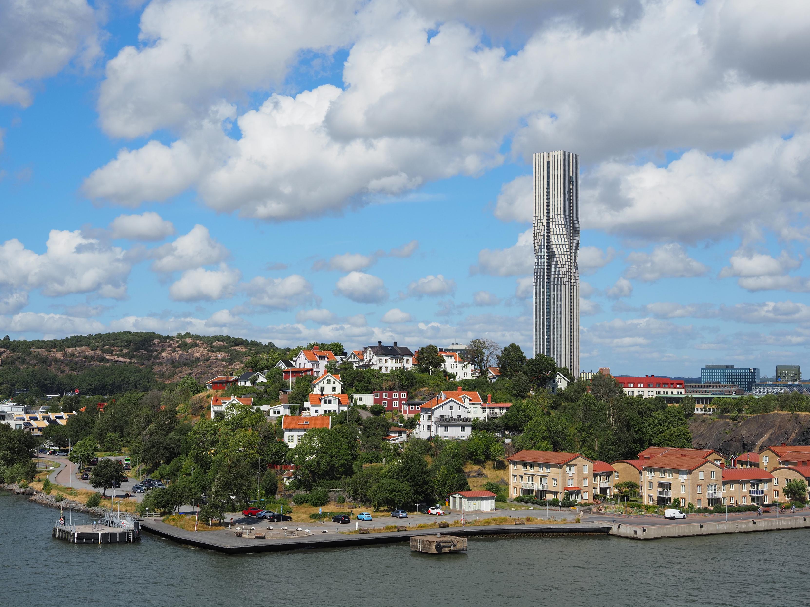 Karlatornet, a white-silver skyscraper with a quadratic footprint consisting of four smaller swquares that are 'twisted' at half of the height, seen from the ferry, trees and buildings in front