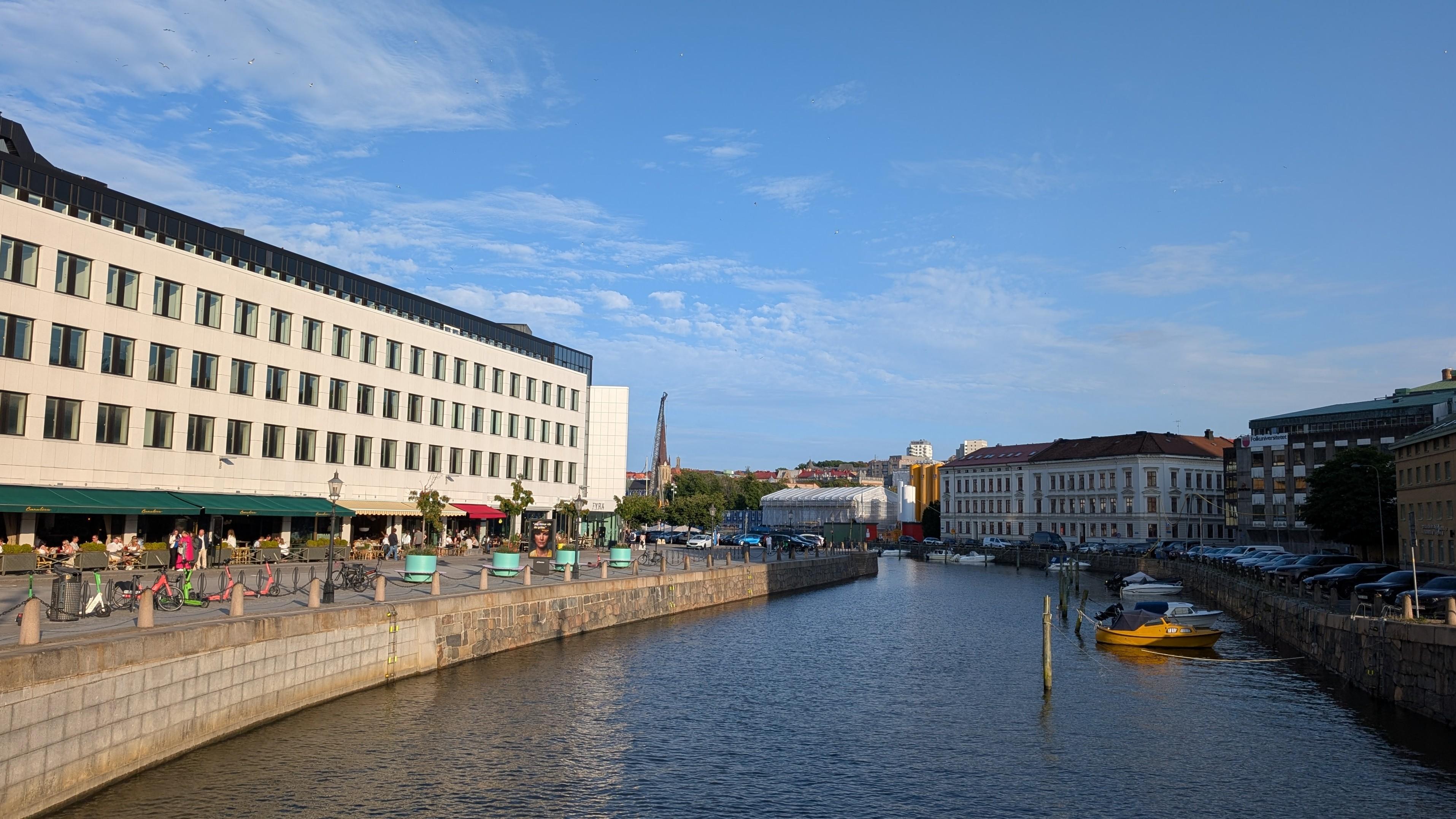 A wide canal aligned with modern buildings with cafés on the left and older buildings on the right side, wide streets and pedestrian zones on both sides, blue sky