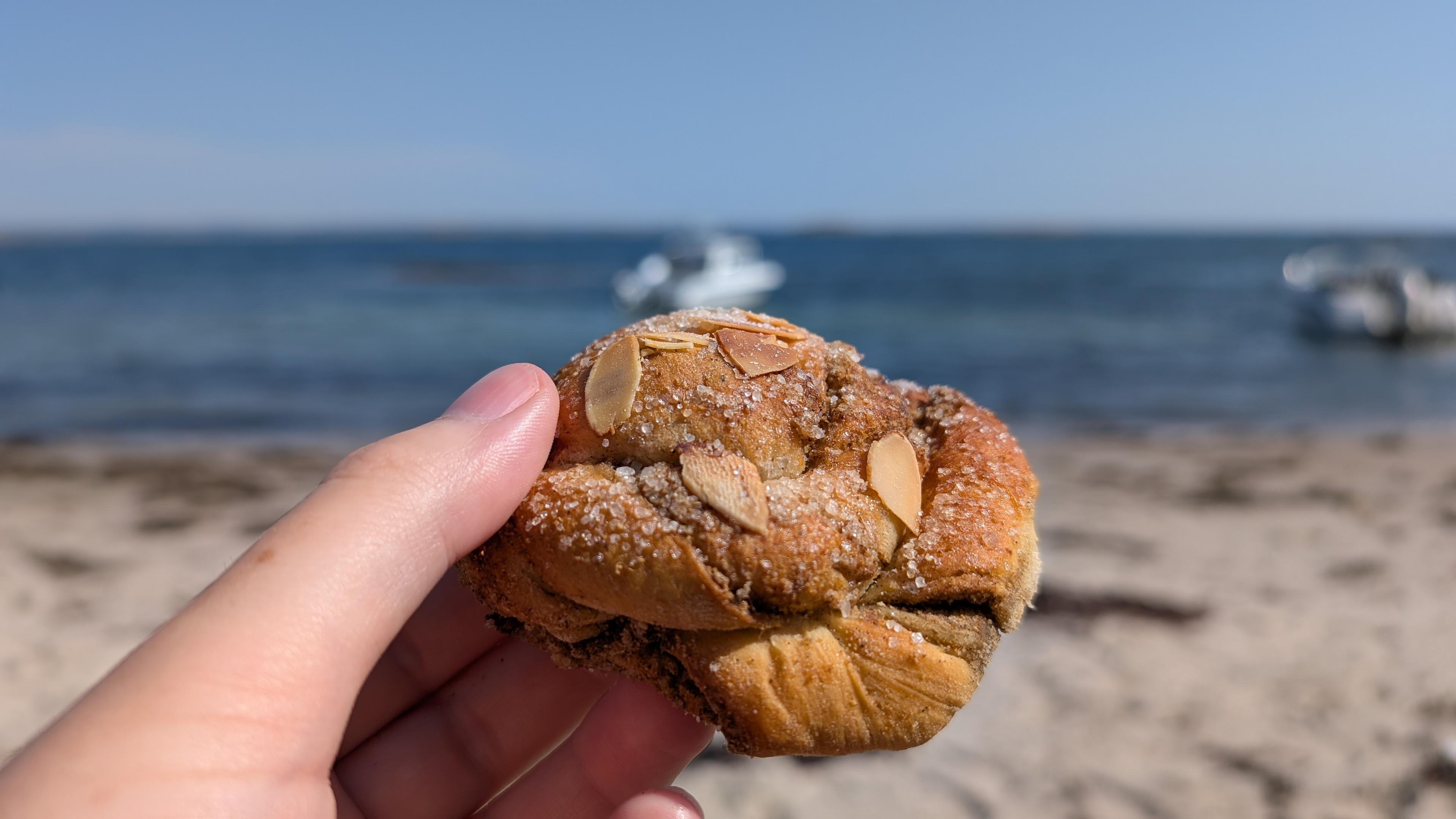 A hand holding a cardamom bun, sand beach and sea with some boats behind it