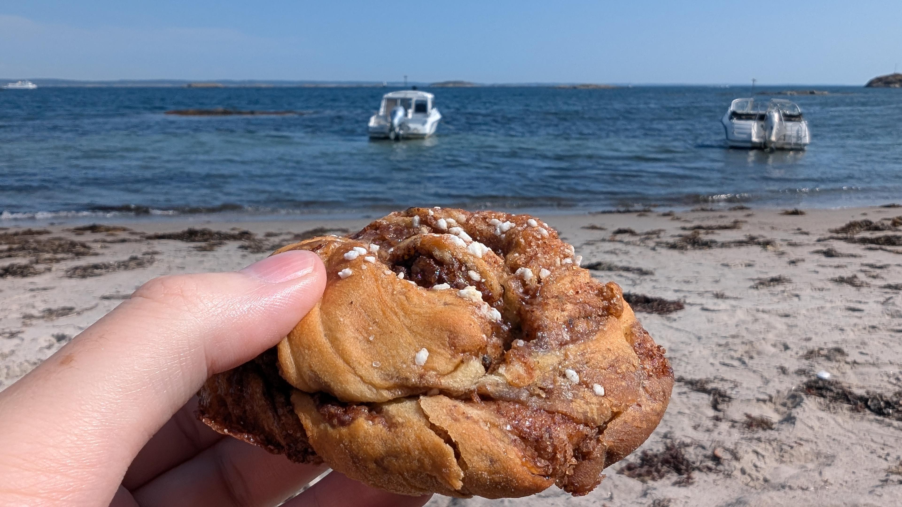A hand holding a cinnamon bun, sand beach and sea with some boats behind it