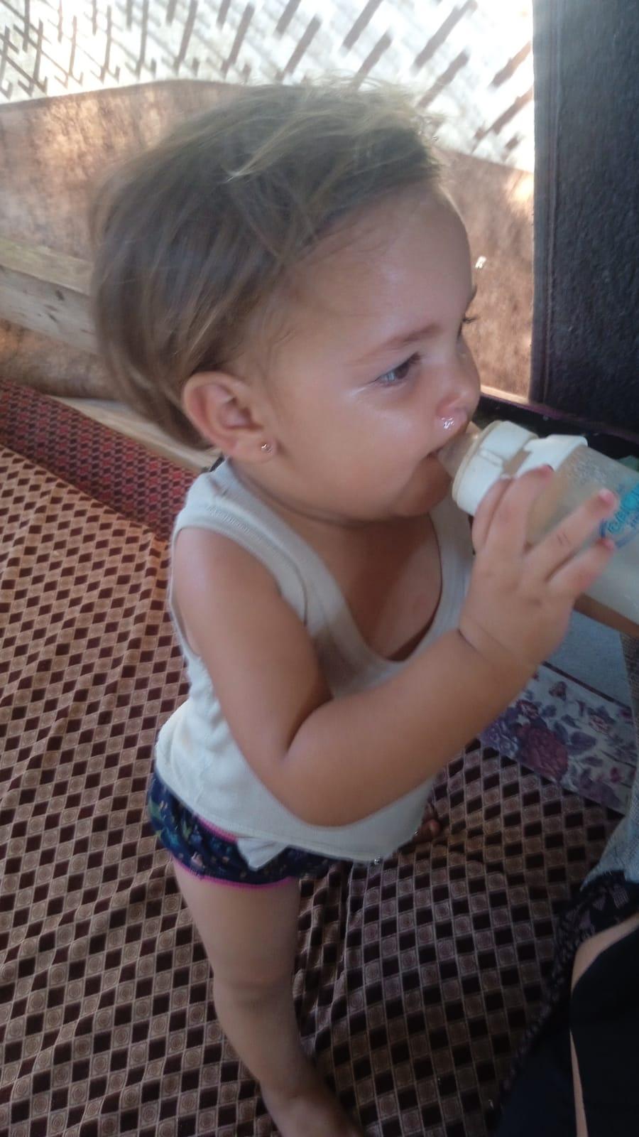 A young child stands in profile, drinking from a white container held with both hands. The child has light brown hair and wears a light-colored tank top and dark patterned shorts. A small earring is visible on their earlobe as they hold the bottle to their mouth. They are standing on a surface covered with a brown and black diamond-patterned fabric. In the background, background, there is a textured wall or fence structure.