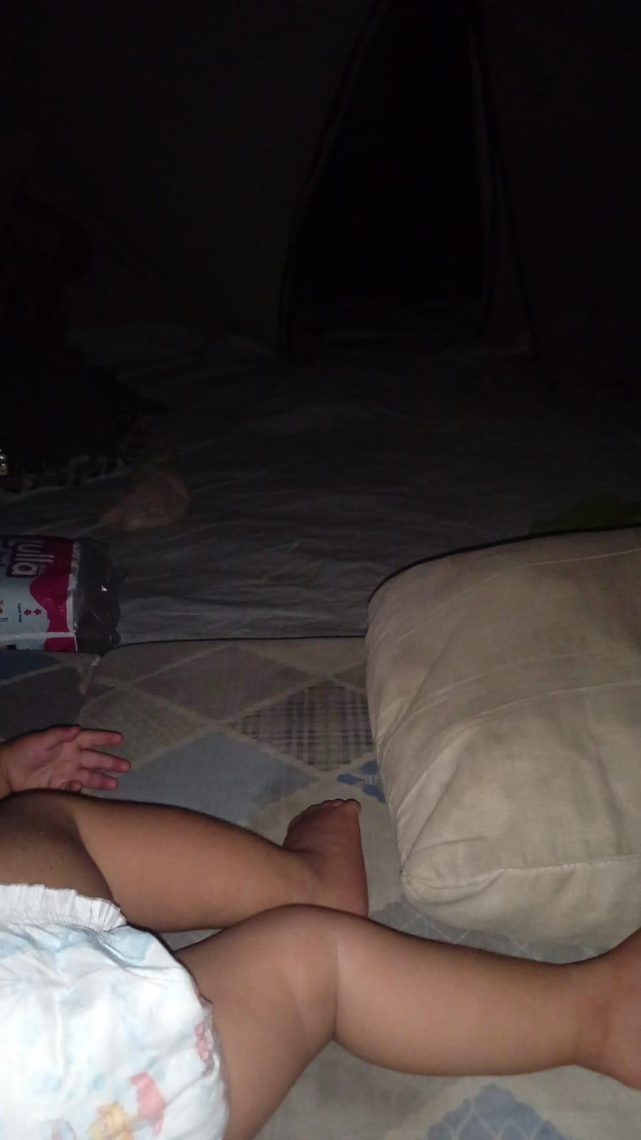 A low-light photograph shows the interior of a dark space with light-colore light-colored fabric flooring and a pointed silhouette in the background. In the foreground, parts of an infant's body are visible, including legs and a hand, wearing a white diaper with a small colorful pattern. One leg is extended toward the right edge of the frame. To the left, there is a pink and white package containing text that includes "ulfa". On the right side of the foreground, a large, beige-colored cushion rests on the floor