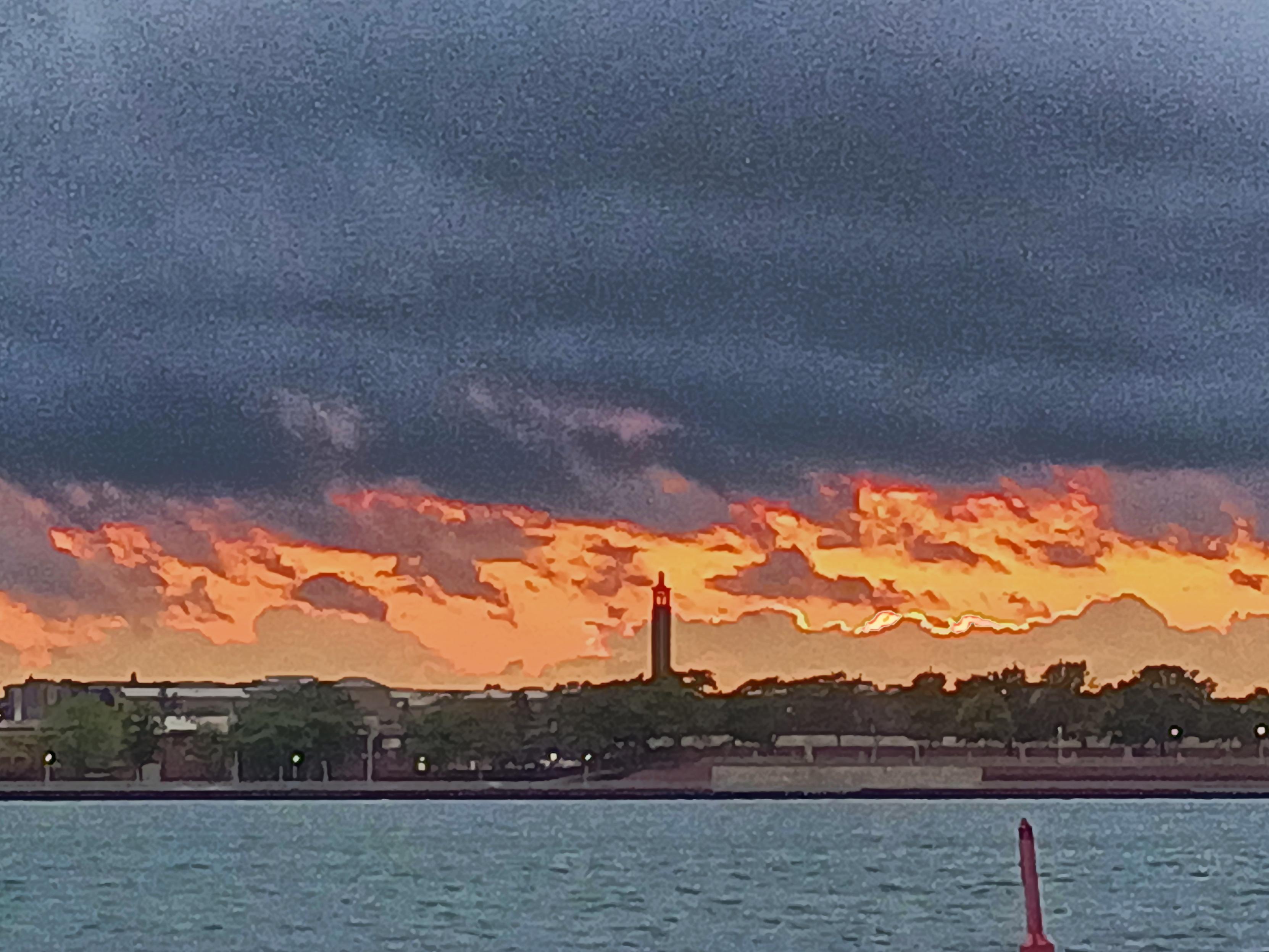 A slightly grainy evening sunset photo with a bright streak of orange below blue dark clouds and a tall spire in the mid ground.