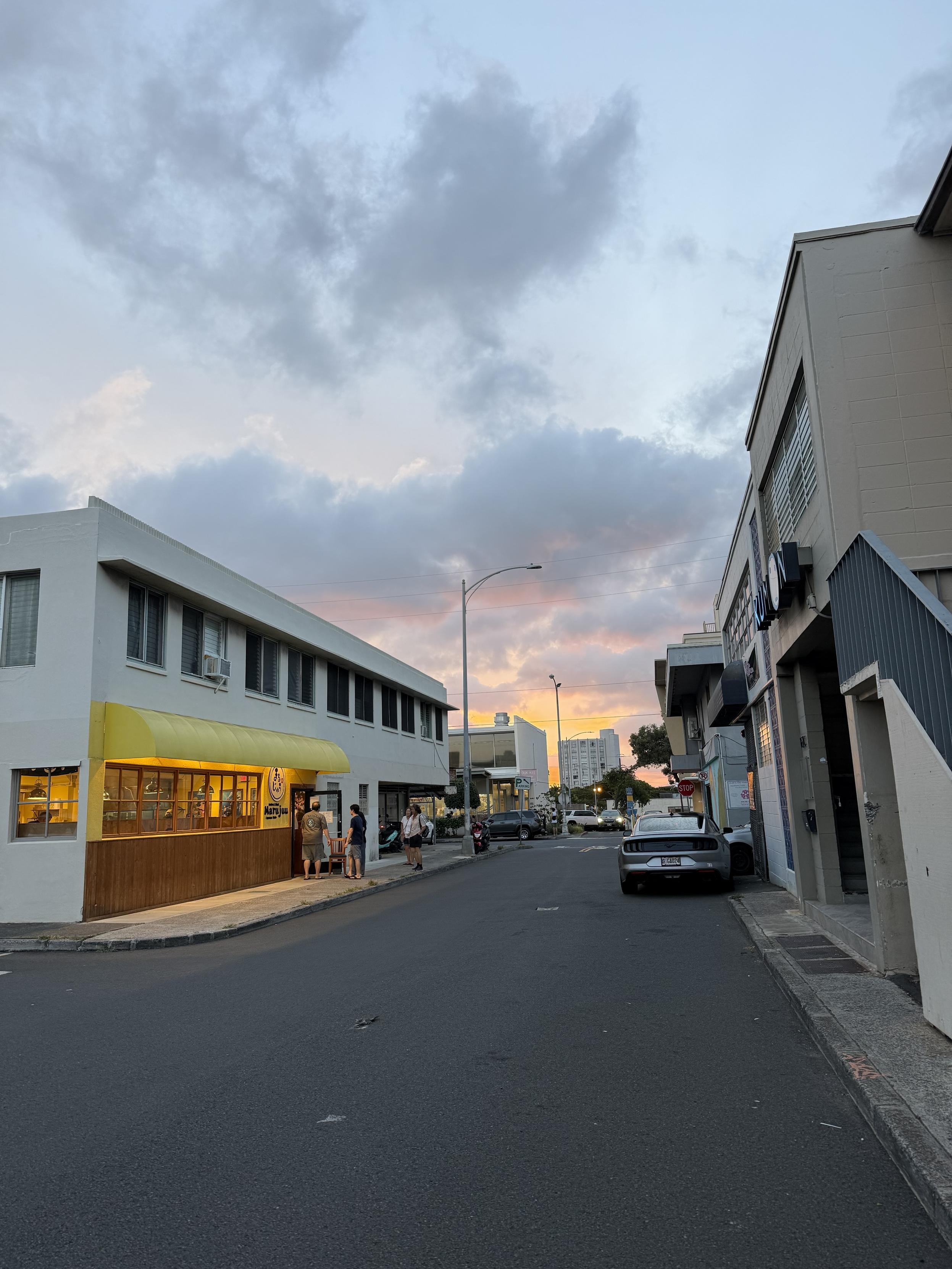 A street scene featuring buildings with a white exterior and a yellow awning. People are gathered outside a shop, and a white car is parked on the side. The sky is colorful with clouds at sunset, casting a warm glow.